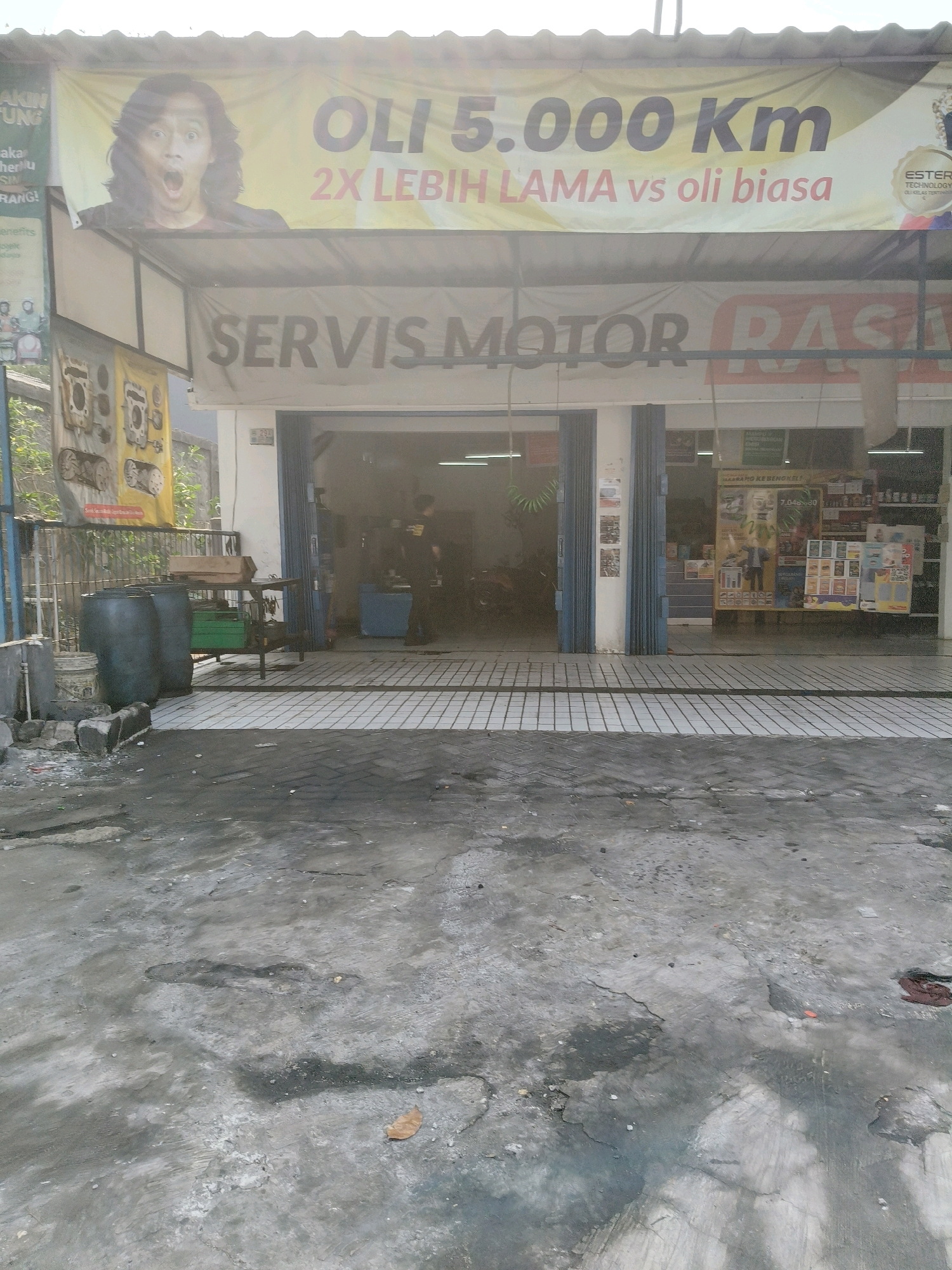 This is a vertical, eye-level, full-shot photograph taken outdoors on a slightly overcast day. The scene depicts the exterior of a motorcycle repair shop, likely located in the city of South Tangerang, Indonesia.
In the foreground, the ground is rough, cracked concrete and asphalt with puddles and oil stains, indicating a well-used and potentially messy area. Two large, dark gray plastic drums are visible on the left side of the image, next to a lighter-colored bucket.
The middle ground shows the entrance to the repair shop. The shop's facade is painted white, with a light gray awning extending outwards. Above the awning, a large yellow banner is stretched across, featuring a surprised-looking person with dark, wavy hair and an open mouth, along with text that reads "OLI 5.000 KM" and "2X LEBIH LAMA vs oli biasa" (meaning "Oil 5,000 KM" and "2X Longer vs ordinary oil"). To the right of this banner, a smaller logo with the word "ESTER" is visible.
Beneath the awning, a gray banner reads "SERVIS MOTOR RASA" (meaning "Motorcycle Service Rasa," where "Rasa" could refer to a brand or style). The shop entrance has blue metal folding doors, partially open to reveal the interior. Inside the shop, a mechanic in a dark shirt is visible, along with a motorcycle and various tools and equipment. To the left of the entrance, a yellow banner hangs with images of engine parts.
In the background, the environment appears to be urban or semi-urban, with some greenery and walls visible beyond the shop. The overall impression is of a functional, working business in an Indonesian city.