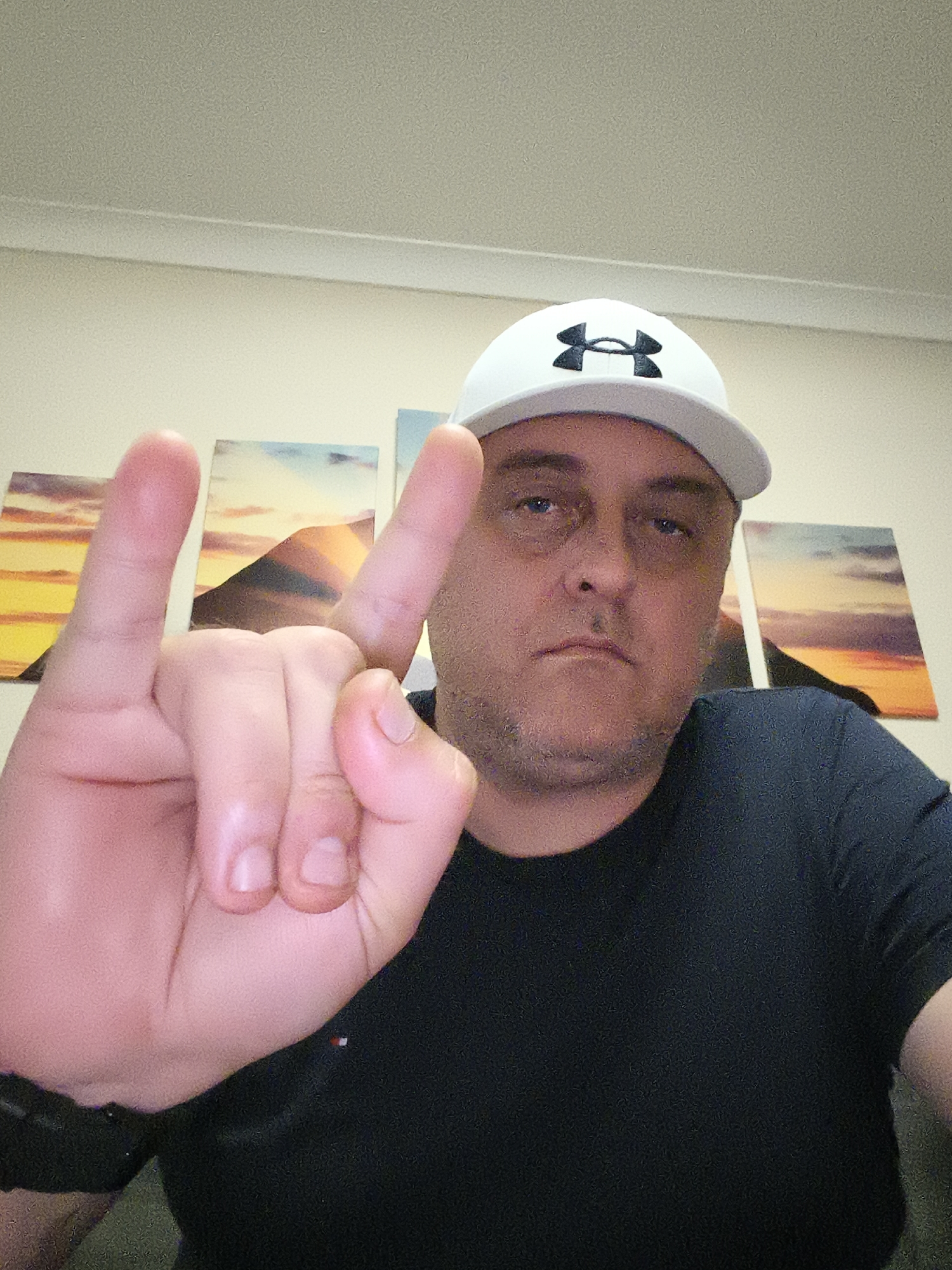 A waist-up portrait features a man indoors, looking directly at the viewer with a neutral to slightly frowning expression. He is wearing a white baseball cap with a prominent black Under Armour logo on the front and a dark (likely black or navy blue) short-sleeved t-shirt. His left hand is raised, performing the "rock on" hand gesture, with the index and pinky fingers extended and the thumb tucked in. The man has short, receding dark hair, visible stubble, and a somewhat flushed complexion. A dark watch or wristband is on his left wrist.
The background shows a light cream-colored wall adorned with three rectangular art prints. These prints depict scenic landscapes, possibly mountains or hills, under a warm-toned sky, characteristic of a sunset or sunrise, featuring hues of orange, yellow, blue, and purple. A plain light-colored ceiling with a molding strip is visible above the wall art. The overall lighting suggests an indoor setting, likely well-lit. The scene is situated in Huntingdonshire, United Kingdom.