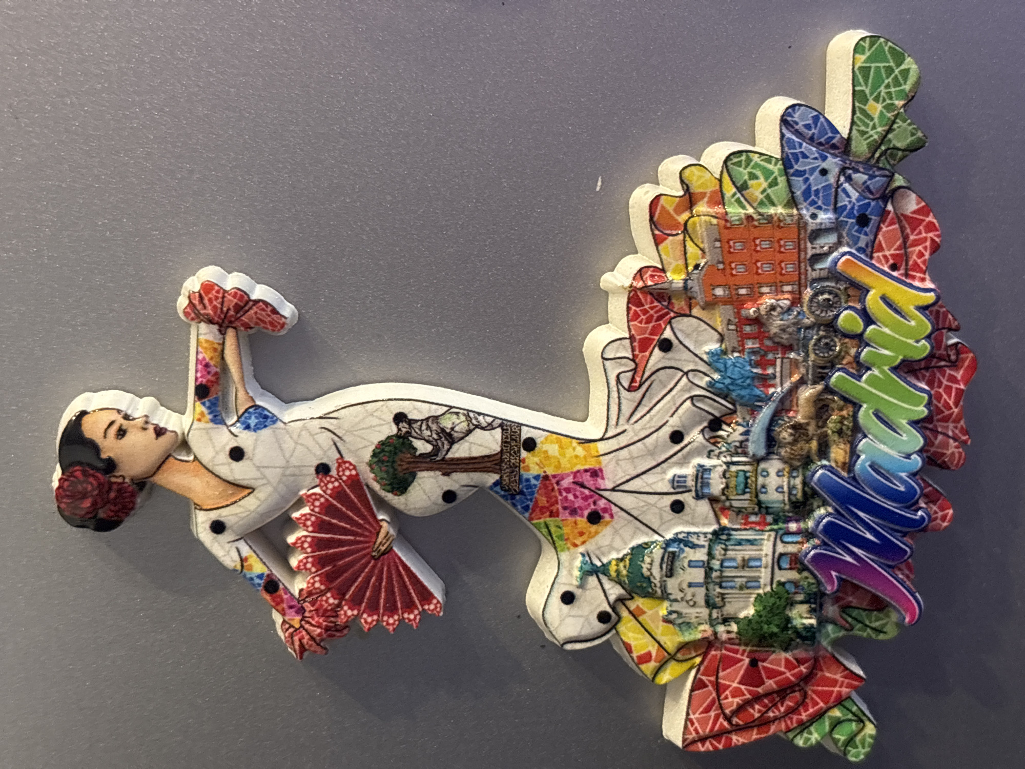 This is a photograph of a souvenir magnet shaped like a flamenco dancer. The dancer is depicted in motion, with one arm raised and holding a red fan, and the other arm bent. She is wearing a white dress with black polka dots and colorful accents. Her hair is dark and styled with a red flower. The body of the dancer is intricately decorated with various colorful patterns, including geometric shapes and images of buildings and trees.
The right side of the magnet features a colorful mosaic of buildings and what appears to be a cityscape. The buildings are detailed with windows and architectural elements. A prominent, multicolored text in a cursive font is visible on the right side, reading "Sofia". The overall impression is that of a vibrant and artistic representation of the city of Sofia, Bulgaria, through the imagery of a flamenco dancer and its iconic landmarks. The lighting suggests a neutral indoor setting, with the magnet positioned against a plain, grey background.