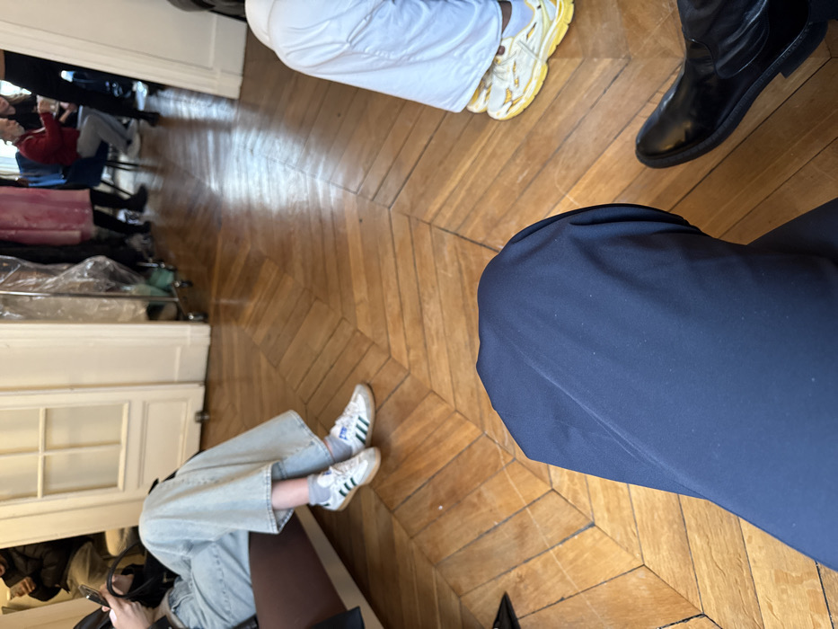 This image depicts an interior scene within a classic Parisian building, identifiable by the distinctive herringbone parquet wood flooring and white-painted double doors. The perspective is from the low vantage point of a seated individual, whose dark blue-clad knee and a polished black leather boot are prominent in the foreground. To the left, another person is seated on a brown upholstered surface, wearing light blue wide-leg denim jeans, grey socks, and white Adidas Samba sneakers with green stripes and gum soles, focused on a smartphone held in their hands. To the right, the lower legs of a third individual are visible, wearing white trousers and yellow, chunky athletic sneakers. Through an open doorway in the background, a deeper room is revealed with multiple figures, including a person in a red sweater seated in a blue chair. Garment racks with various clothing items, some covered in clear plastic, are also discernible, suggesting a fashion-related context such as a showroom, fitting area, or backstage preparation space within the city of Paris, France.