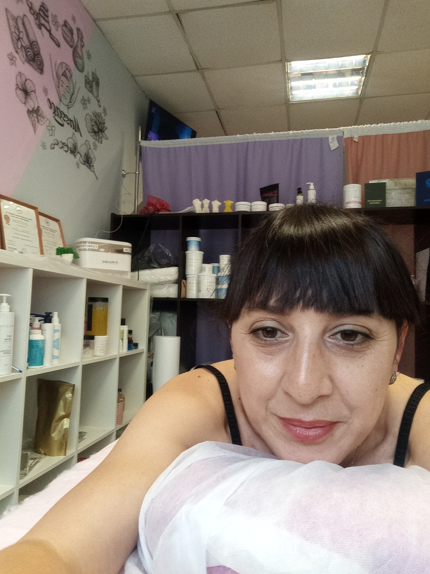 The image shows a woman with dark hair and bangs, wearing a black tank top, lying on a pink surface covered with a white sheet. She is looking towards the camera with a slight smile. The background reveals a room with shelves filled with various products, likely in a spa or salon setting. The shelves contain bottles, jars, and containers of different sizes and colors. On the wall, there is a mural with a floral design and some lettering. A television screen is visible in the upper left corner. The ceiling has fluorescent lighting panels. The scene suggests an indoor environment, possibly a treatment room.