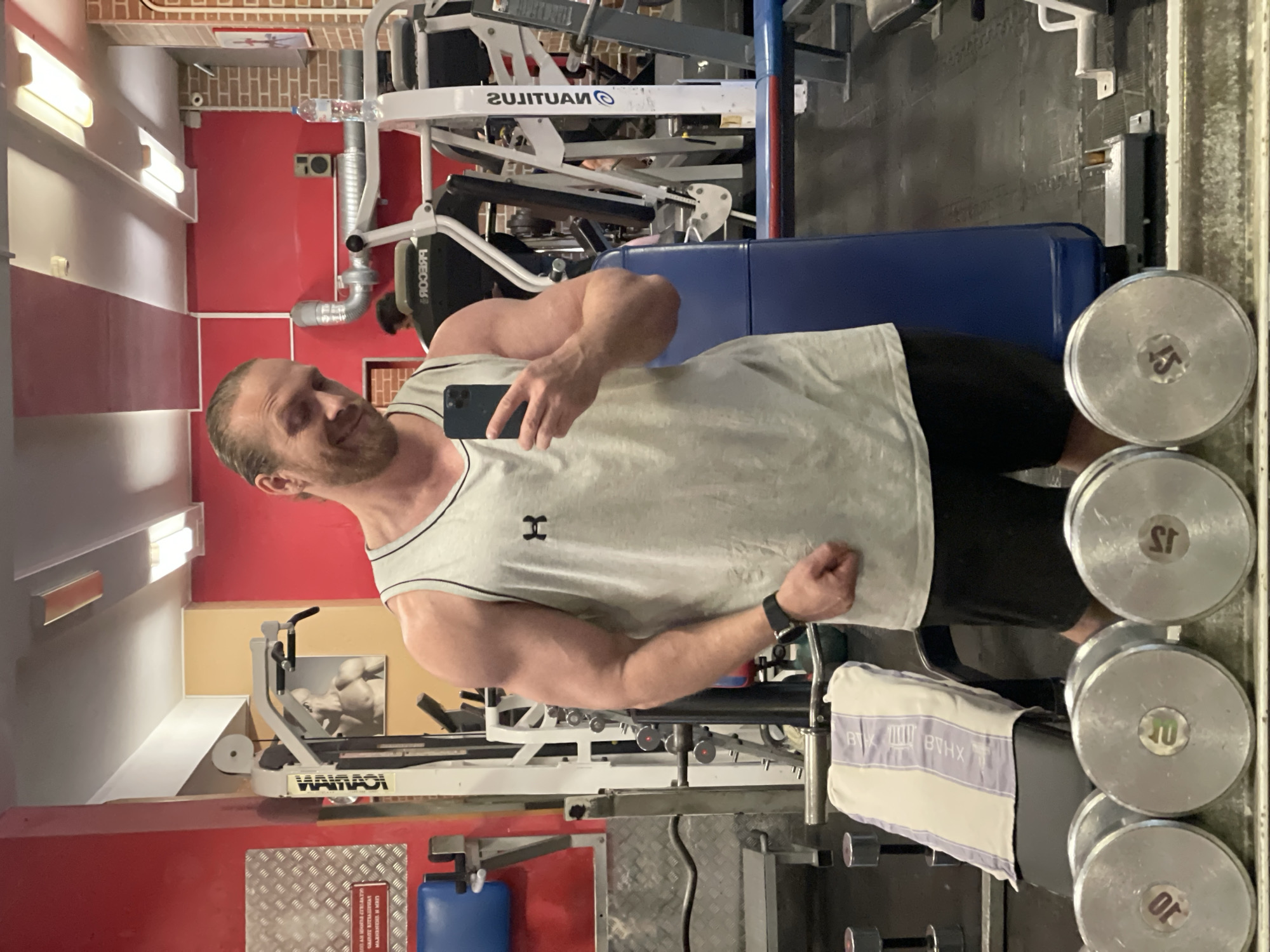 A fair-skinned man with blonde hair pulled back in a ponytail, wearing a light gray Under Armour tank top and black shorts, poses for a selfie in a gym. He has a smile on his face and is holding a black smartphone in his right hand. In the background, there are various weightlifting machines, dumbbells, and a bench with a towel. The gym walls are painted red and yellow, and there are exposed pipes and lighting fixtures on the ceiling. The overall environment suggests a typical gym setting in Saint Petersburg, Russia.