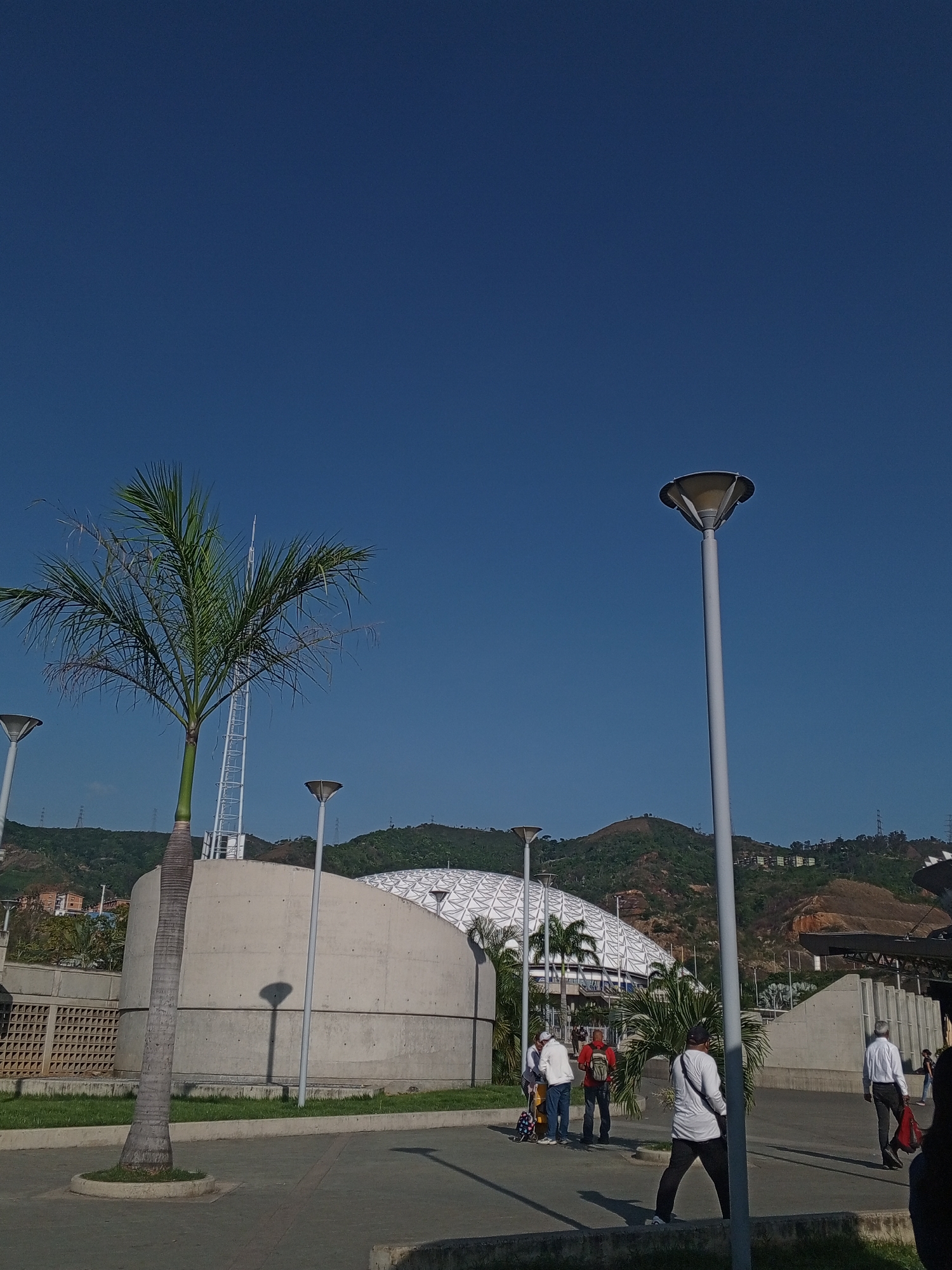 The image shows an outdoor scene in Caracas, Venezuela, featuring a tall palm tree, several modern-style streetlights, a curved concrete building (possibly a stadium or convention center), and a backdrop of green hills. People are present, some standing and conversing, others walking along the paved area. The sky is a clear, deep blue. The scene appears to be taken during the day with strong sunlight and long shadows.