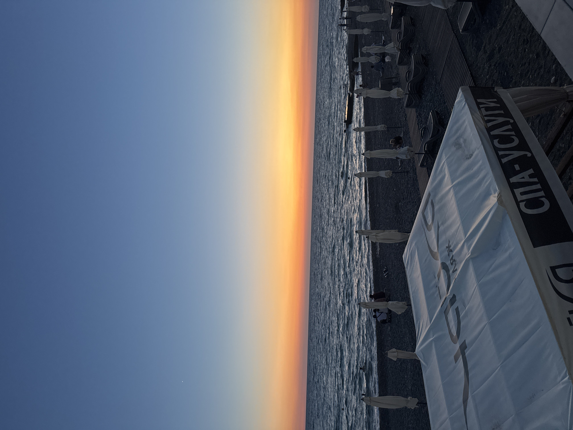 This is a wide-angle, low-angle shot of a beach in Sochi, Russia, during sunset. The sky transitions from a deep blue at the top to a vibrant orange and yellow band just above the horizon. The sea is visible in the mid-ground, with gentle waves and a few people swimming or wading. The foreground is dominated by a beach with dark pebbles, lined with closed umbrellas and lounge chairs. In the immediate foreground, a white tent with Cyrillic text reading "СПА-УСЛУГИ" (Spa Services) is partially visible. The overall atmosphere is calm and serene, typical of a beach at dusk.