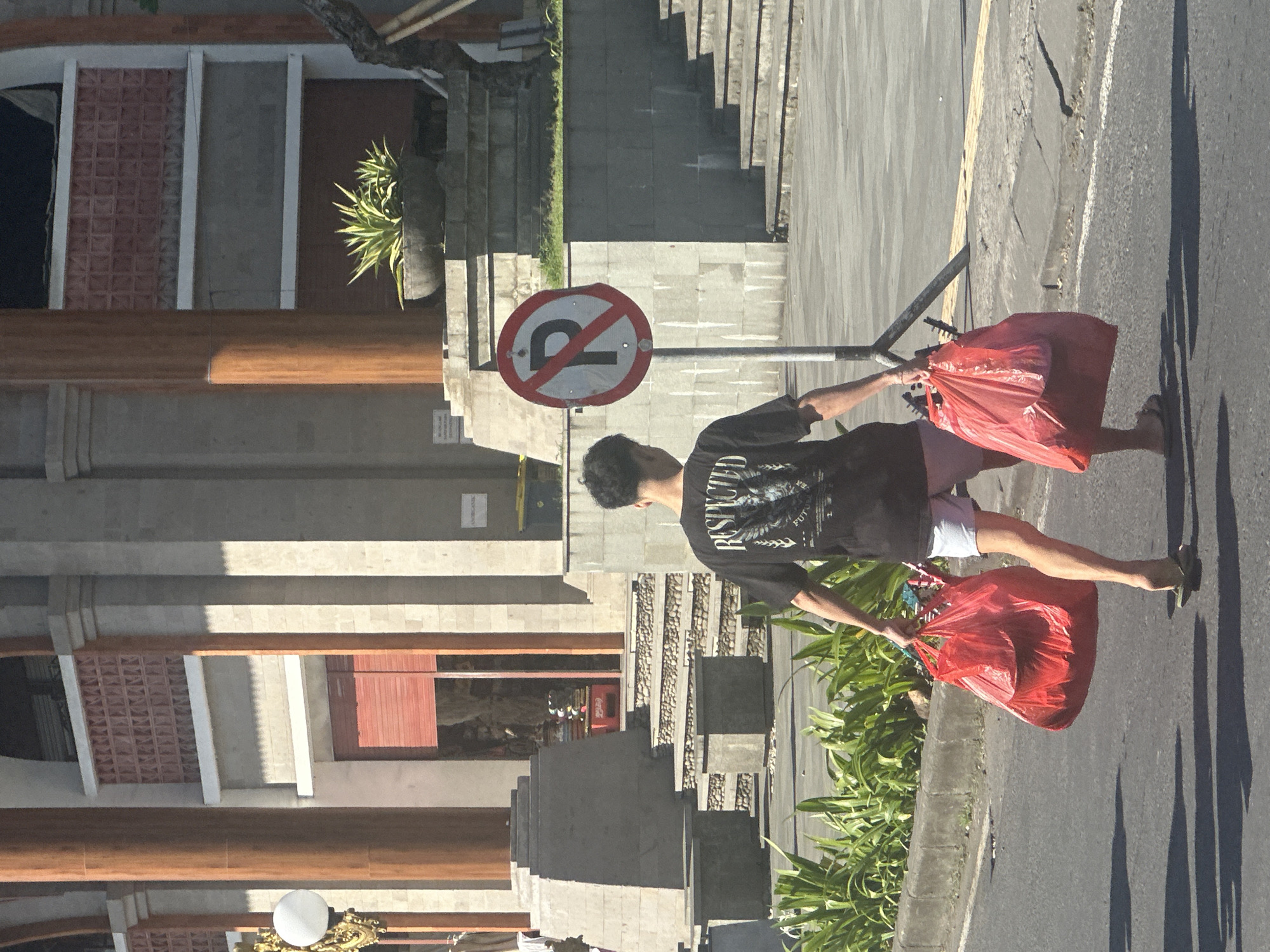 A young man with dark hair walks away from the camera down a street in Ubud District, Indonesia. He is wearing a black t-shirt with a graphic design and light pink shorts, and flip-flops on his feet. In his left hand, he carries a large red plastic bag filled with items. In his right hand, he carries a similar, slightly smaller red plastic bag. To his left, a "no parking" sign is visible on a metal pole. Behind him, a large building with columns and decorative brickwork can be seen, with steps leading up to it. Lush green plants line the edge of the street. The sun is casting long shadows on the asphalt.