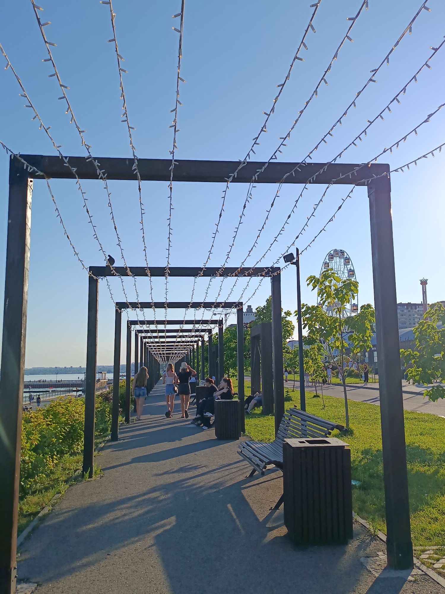 The image captures a sunny day at a park in Saratov, Russia. A pathway is lined with a pergola structure adorned with strings of small lights, creating a charming effect. Several people are enjoying the outdoors. In the foreground, three young women are walking away from the camera along the path, dressed casually for warm weather. To their right, two individuals are seated on a park bench, appearing relaxed. Further down the path, other people can be seen strolling or sitting, suggesting a leisurely atmosphere.
In the background, a Ferris wheel stands tall against a clear blue sky, indicating a recreational area or amusement park nearby. Green grass and trees line the pathway, contributing to the park-like setting. A body of water, likely a river, is visible to the left, with a distant cityscape on the horizon. The lighting suggests it is either late afternoon or early morning, with the sun casting long shadows. The overall mood is peaceful and inviting, conveying a pleasant day out in an urban park. There is no visible text in the image.