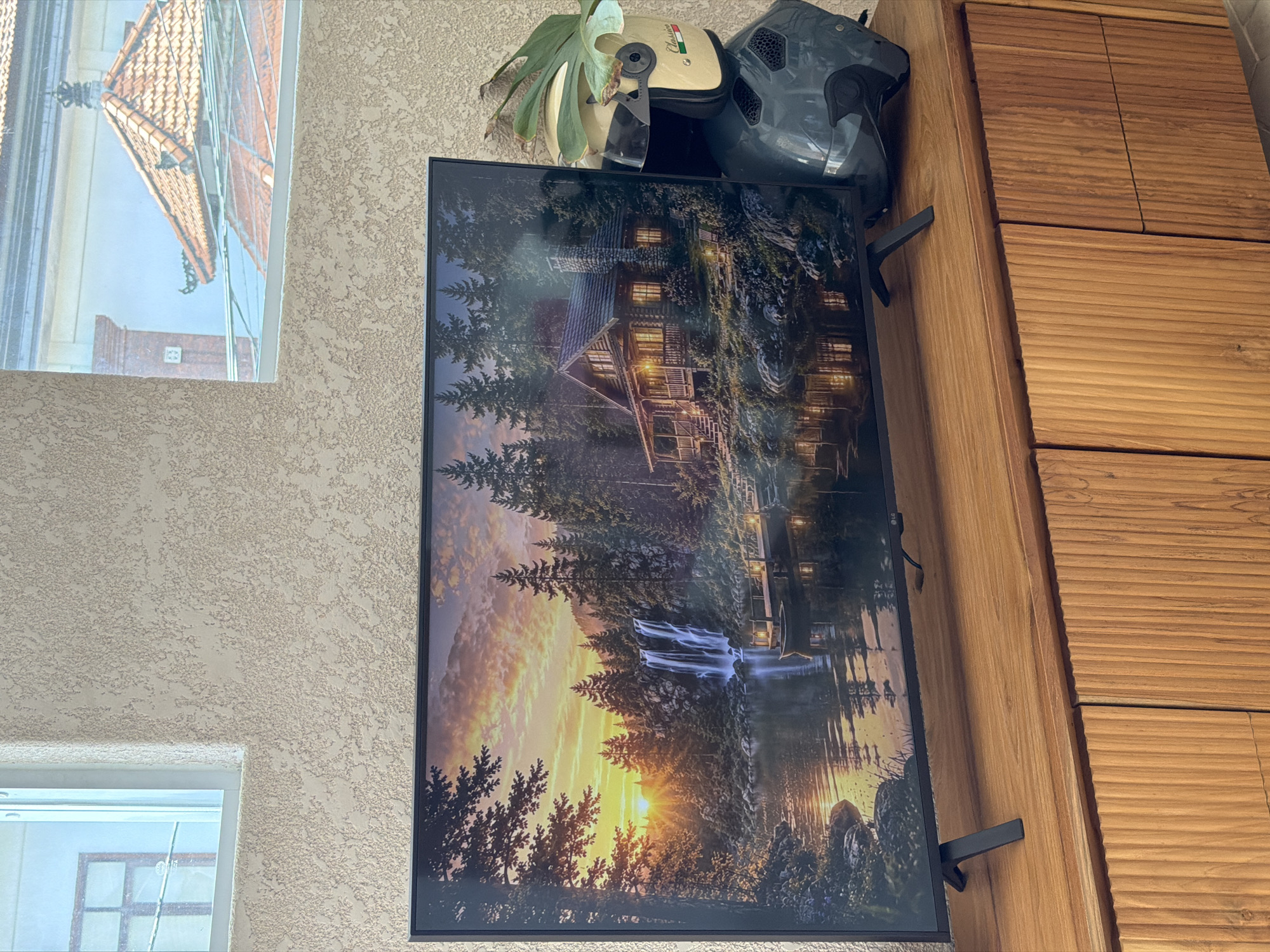 The image shows a flat screen television mounted on a wooden cabinet. The screen displays a nature scene with a lake, waterfall, and a cabin. On top of the television, there is a staghorn fern, a cream-colored motorcycle helmet, and a gray motorcycle helmet. A window is visible on the left side of the image, reflecting a building with a tiled roof. The wall behind the television and window has a textured, off-white finish. The scene appears to be indoors. There is no direct evidence to confirm the location is Mengwi, Indonesia.