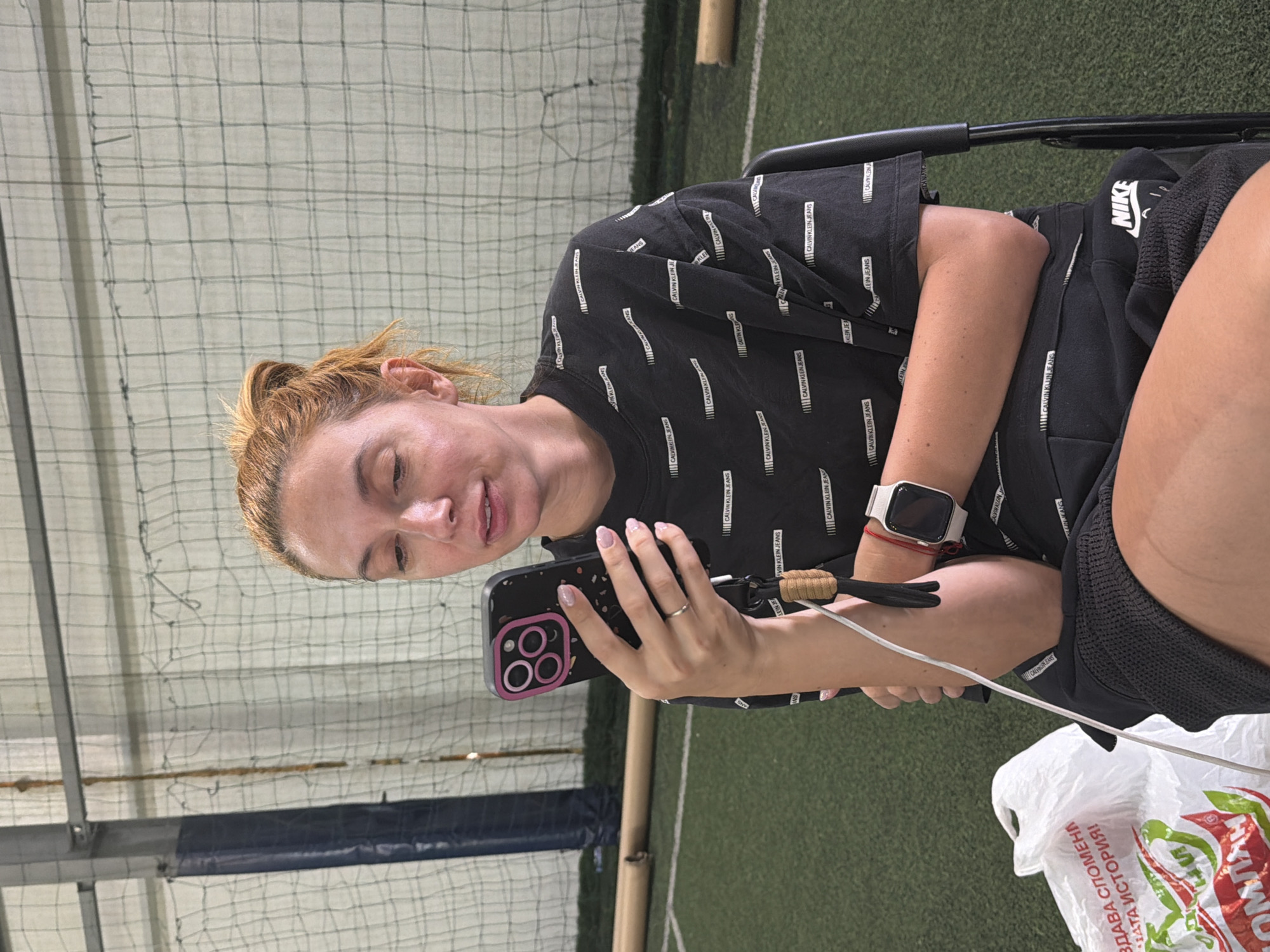 A woman with auburn hair pulled up is seated indoors, holding a smartphone in her left hand and looking at it. She is wearing a black t-shirt with a repeating white Calvin Klein Jeans logo pattern, black shorts, and a white smartwatch on her left wrist. A white plastic shopping bag with red and green text is to her left, and a black chair is behind her. The background features a green turf-like floor, a net on the wall, and metal structures, suggesting an indoor sports facility. The image does not contain any specific landmarks or indicators of a location within Sofia, Bulgaria.