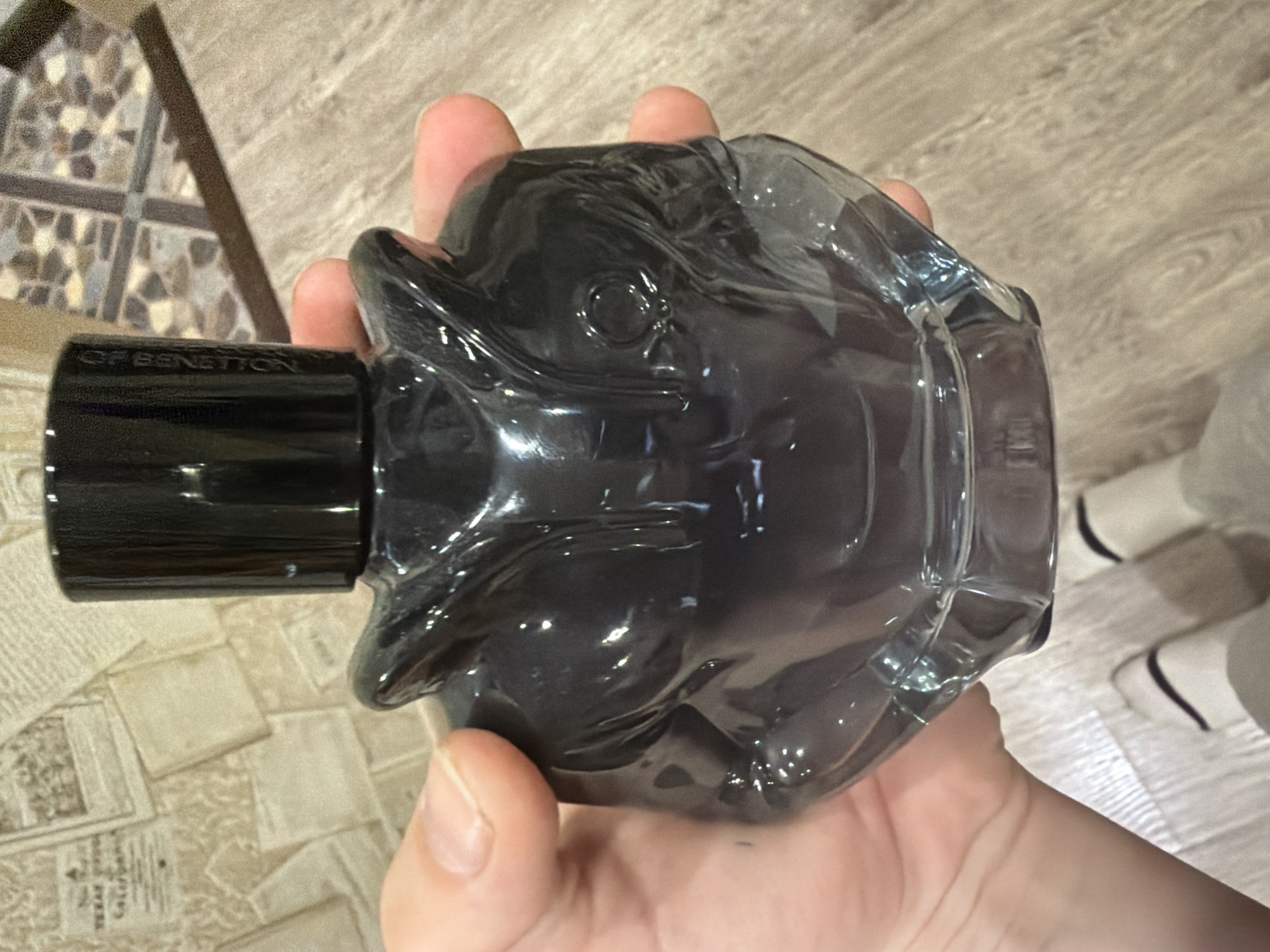 A person holds a bottle of "United Colors of Benetton" perfume, its black cap and translucent body shaped like a cartoonish head. The background shows a mosaic-patterned surface and a light-wood-grain surface; in the bottom right, white objects with dark trim are visible. It is impossible to tell with certainty if this photo was taken in Zaprudnya, Russia.