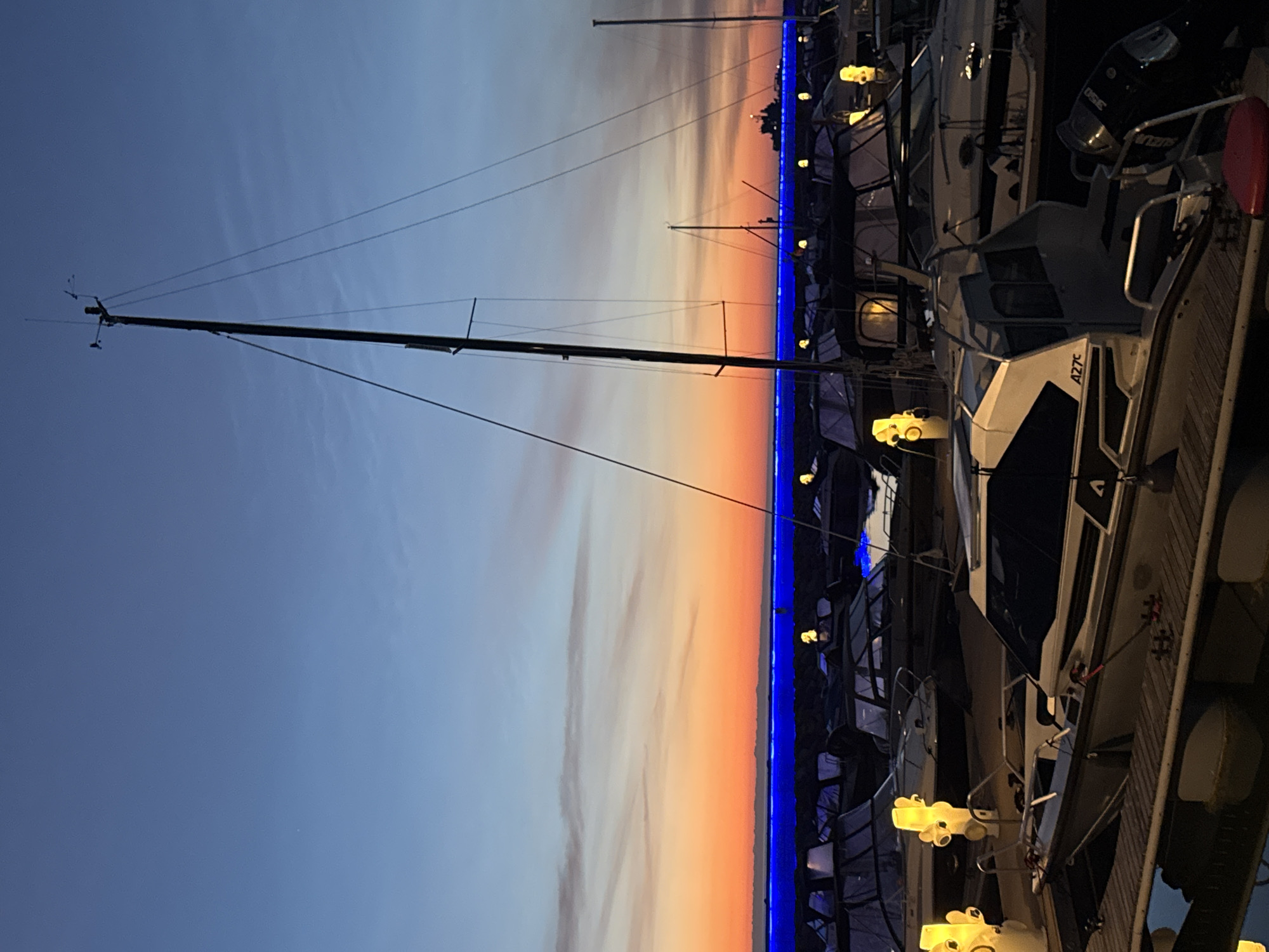 This image depicts a harbor in Tallinn, Estonia, during twilight. The sky transitions from a deep blue overhead to vibrant orange and pink hues near the horizon, with thin clouds streaking across. A bright blue light strip runs horizontally across the background. The foreground features several docked boats of various sizes, many covered with tarps. A prominent sailboat with a tall mast stands in the middle ground, its rigging silhouetted against the sky. Several small, yellow, abstract sculptures are placed along the docks, some near the boats. A distant ship is visible on the water. The scene is illuminated by the fading daylight and artificial lights from the sculptures and the blue light strip.