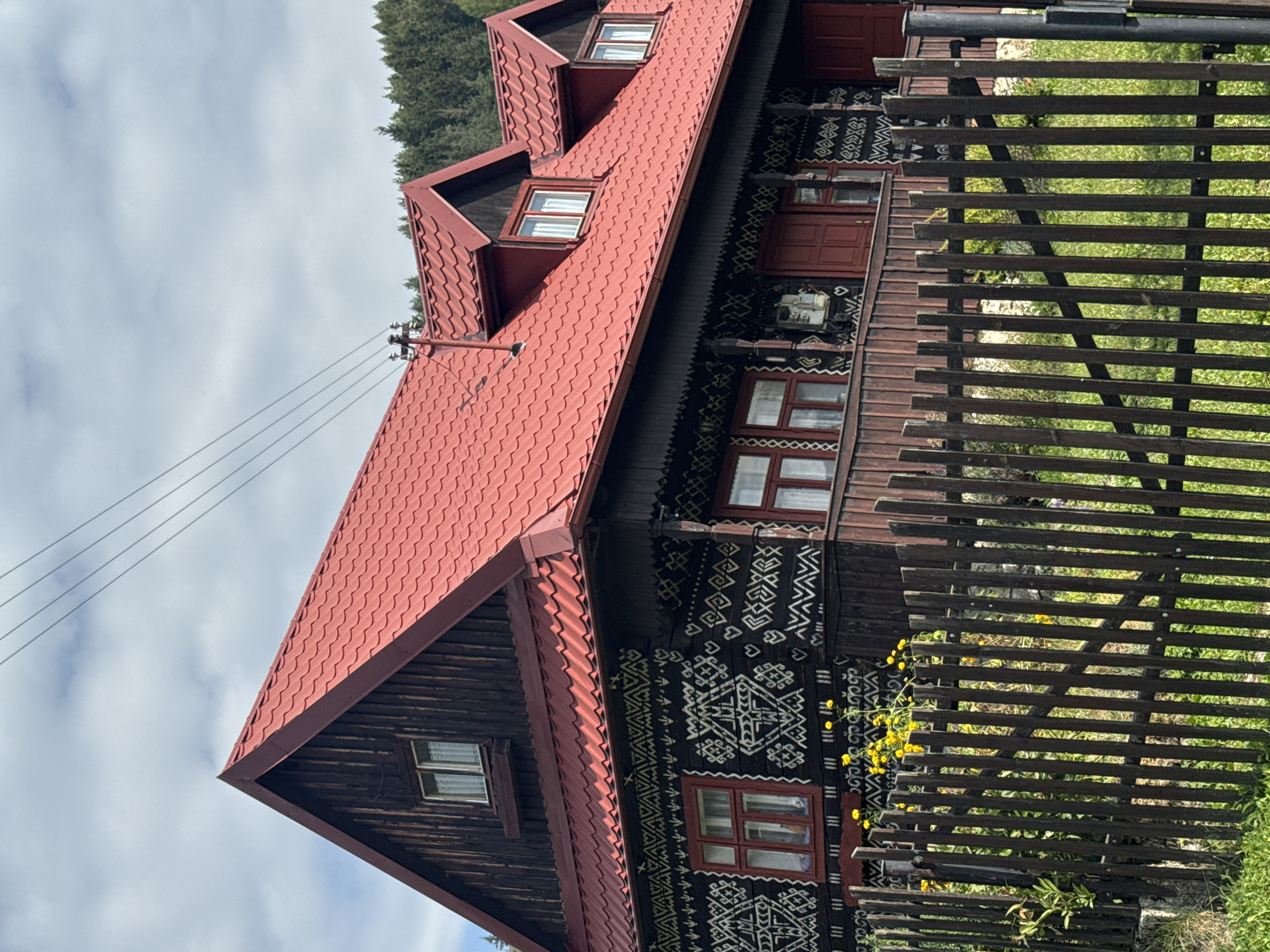 The image shows a traditional wooden house in the village of Čičmany, Slovakia. The house has a distinctive red tiled roof and dark wooden walls adorned with intricate white folk patterns. There are several windows on the facade, some with red frames. A wooden fence encloses the front of the property, with green grass and yellow flowers visible behind it. The house is situated in a rural setting, with a forest visible in the background. The sky is partly cloudy.