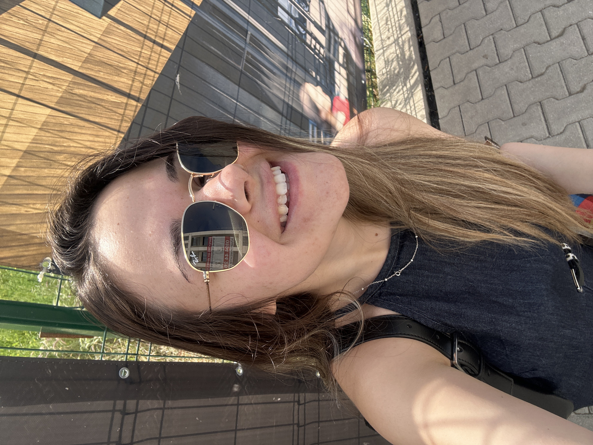 An outdoor selfie captures a young adult female with long, light brown hair, smiling broadly with her teeth showing. Her head is tilted significantly to the right, and the image itself appears to be rotated 90 degrees clockwise. She wears gold-framed Ray-Ban sunglasses, which prominently reflect a building with dark windows and what appears to be Cyrillic text, possibly from a sign, in the lenses (e.g., "ЗА АДРЕС" and other fragmented words). She is dressed in a dark, possibly denim, top or dress, adorned with a thin silver necklace. A black strap or belt is visible near her right shoulder/arm.
The background, viewed from the rotated perspective, features a modern building facade with horizontal elements and strong shadows on the upper left, and grey interlocking concrete pavers (a sidewalk or pathway) on the right. A green metal fence is visible in the lower left portion of the image. The bright sunlight indicates a clear, sunny day, likely midday or afternoon, casting distinct shadows. The overall scene suggests an urban setting in Sofia, Bulgaria, characterized by architectural elements and public spaces. Her wide, joyful smile conveys a cheerful emotion.