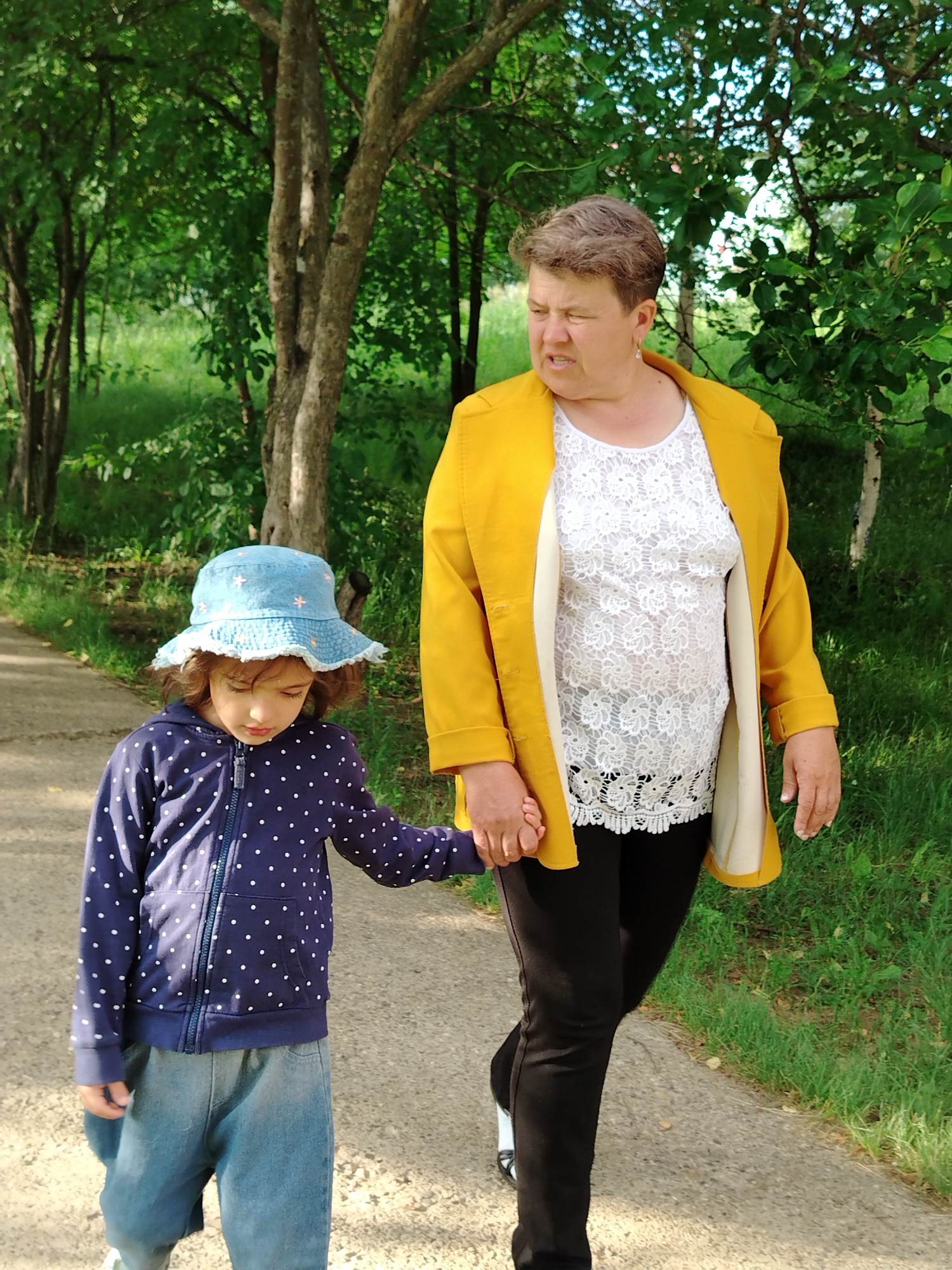 The image depicts a woman and a young child walking hand-in-hand on a paved path in a park-like setting. The woman, on the right, is wearing a bright yellow coat over a white lace top and black pants. She has short, light brown hair and is looking to her left with a concerned expression. The child, on the left, is wearing a blue polka dot hoodie, loose-fitting denim pants, and a light blue sun hat with a ruffled brim. The child is looking down. The background consists of lush green trees and grass, suggesting a natural or park environment. The lighting indicates it is daytime. The scene is likely outdoors in a park, common in towns like Noviy, Russia, where residents would utilize such public spaces for recreation. The interaction between the two individuals appears to be one of guidance and protection, with the woman leading the child.
