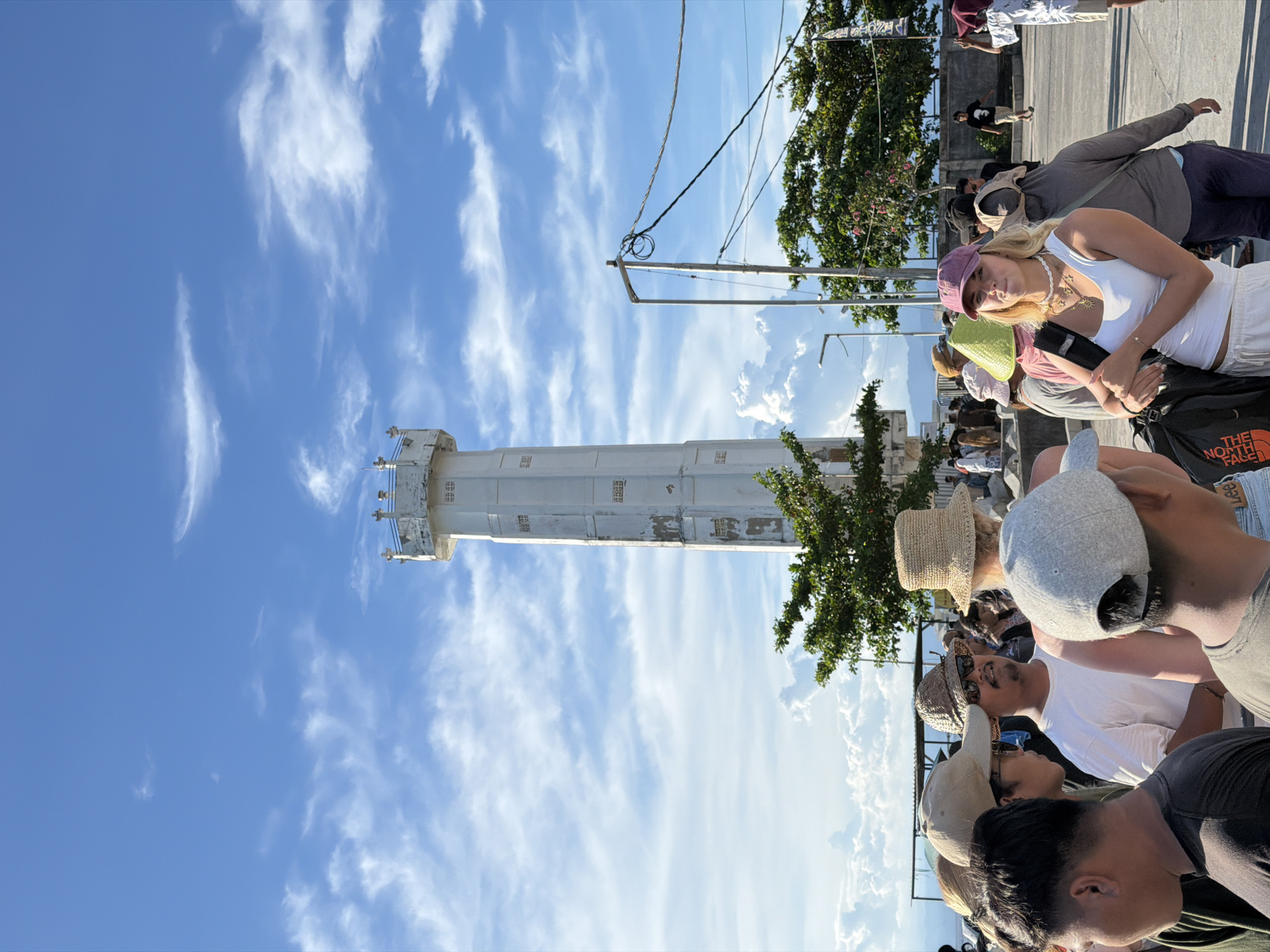 The image captures a bustling outdoor scene on a bright, sunny day, featuring a tall lighthouse and a crowd of people. The photo is taken from a low angle, looking up towards the sky and the top of the lighthouse.
In the midground, a prominent, cylindrical white lighthouse stands tall against a vibrant blue sky dotted with scattered white, wispy clouds. The lighthouse appears to be made of concrete or a similar material, showing some signs of weathering. Above the lighthouse, power lines and utility poles crisscross the sky.
A diverse group of people, likely tourists, fills the foreground and midground. In the midground, a young woman wearing a white tank top, light-colored shorts, and a purple baseball cap looks directly at the camera. She has a gold necklace and carries a black backpack with "THE NORTH FACE" logo clearly visible on it. Around her, other individuals are seen from various angles, many wearing hats (including straw hats and baseball caps) and casual clothing, suggesting a warm climate. Some people are looking around, while others are seen from behind.
Trees with green foliage are visible to the right of the lighthouse and in the background, adding to the natural setting. The ground appears to be paved. The bright sunlight indicates it is daytime, and the clear sky suggests good weather. The overall atmosphere is lively, characteristic of a popular tourist destination. No other significant text is clearly legible in the image.