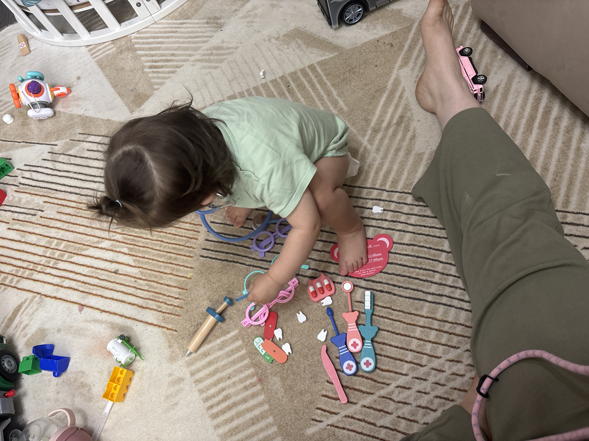 A child plays with a toy doctor's kit on a carpeted floor in a room that also contains a baby's crib and a person's legs. The child, wearing a light green shirt, is seated on the floor and surrounded by various toy medical instruments, including a stethoscope, a syringe, and several dental tools. Next to the child is a person's legs, clad in olive green pants, with a pink toy truck placed near the person's bare foot. Other toys, like trucks and blocks, are scattered around the floor. This image was taken in an indoor setting, specifically within a residence located in the city of Chaikovsky, Russia.