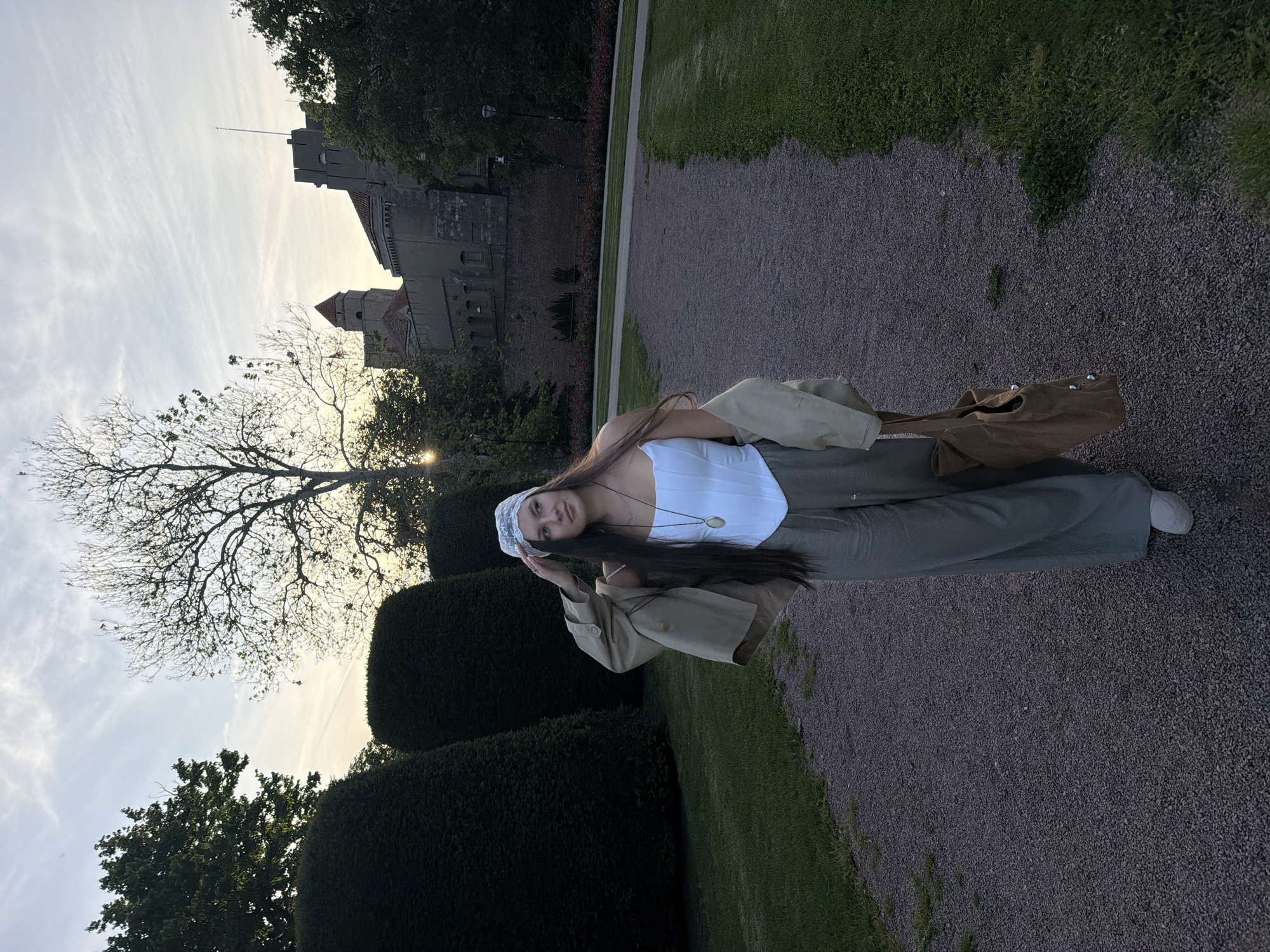 A young woman stands in a park. She wears a white top, olive-green pants, a white bandana, and a tan coat. She holds her hand up to her head. A brown bag lies on the ground behind her. The ground is gravel, bordered by green grass and manicured hedges. In the background is a stone castle and a tree with bare branches against a bright sky. The location is not Belgrade, Serbia.