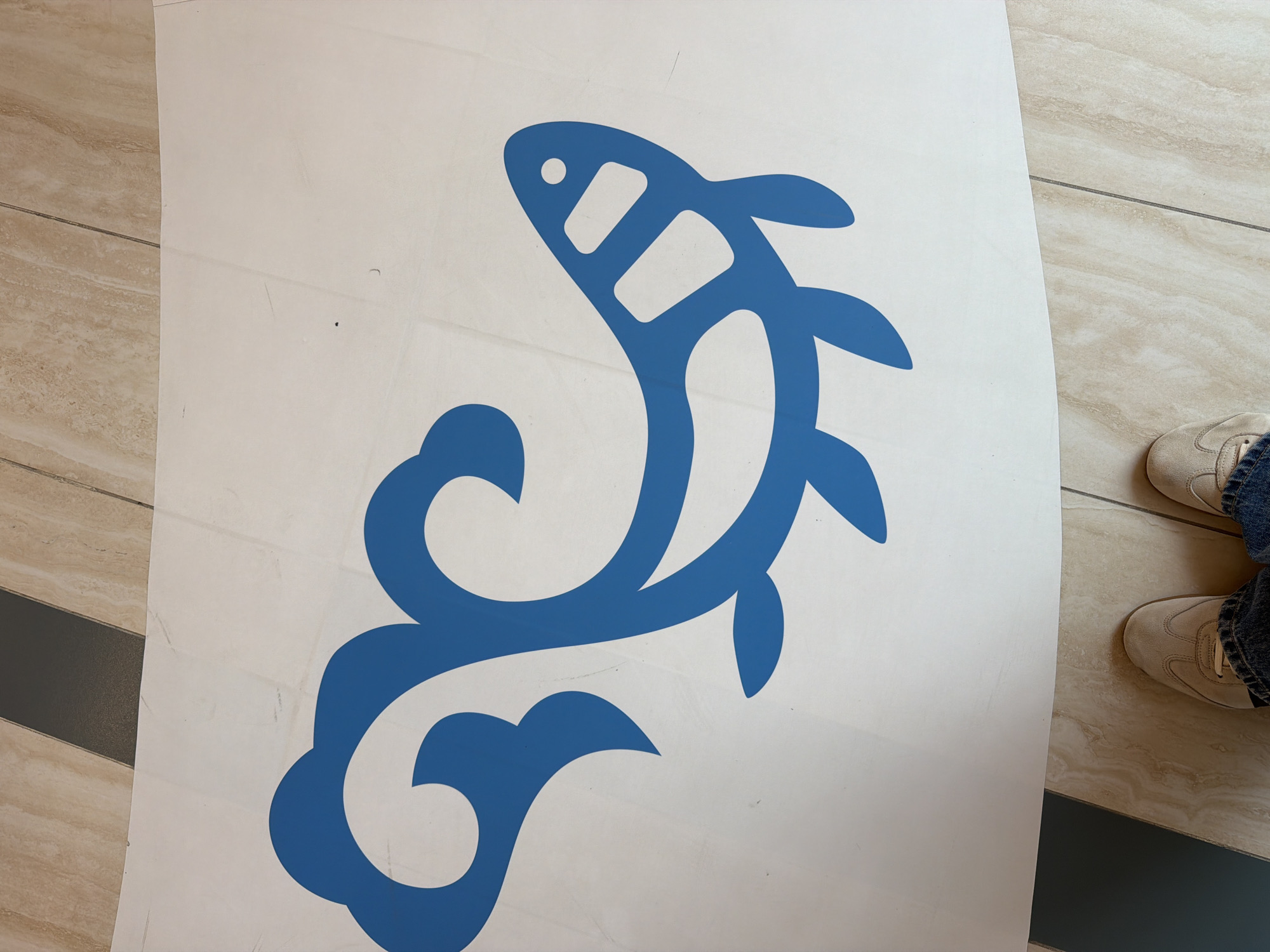 A blue stylized aquatic creature, resembling a fish or a whale, is adhered to a large white sheet, possibly a sticker or decal. The creature has an abstract, flowing design with rounded fins and tail, and distinct rectangular markings along its body. The white sheet is laid out on a floor tiled with a neutral, light beige marble pattern. At the bottom of the image, the feet of a person wearing beige sneakers and blue jeans are visible, suggesting the sheet is on the floor. The background features a strip of darker flooring, possibly slate or a dark tile, running parallel to the marble tiles. There are no discernible Yuzhno-Sakhalinsk landmarks or indicators present in the image.