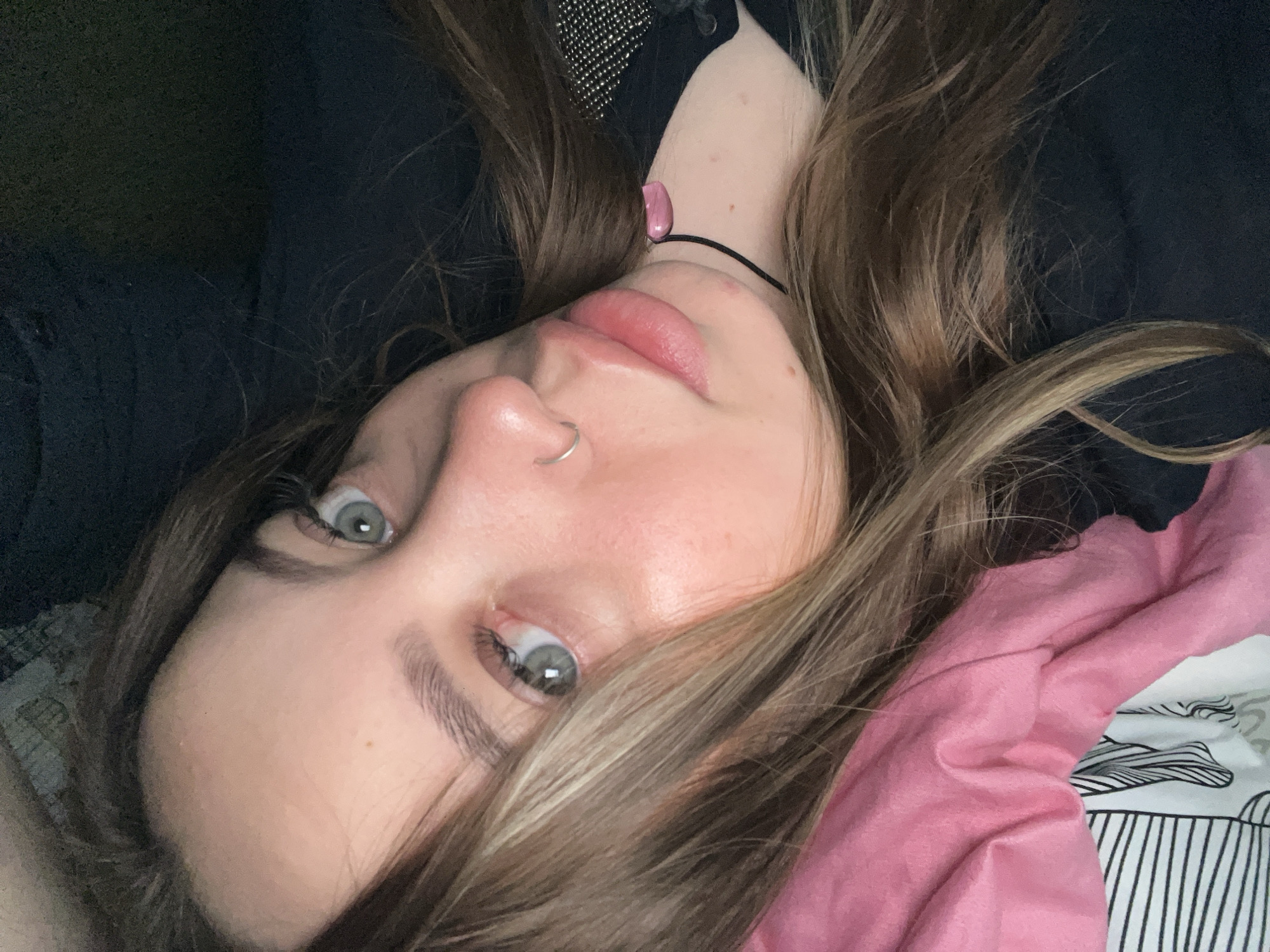 A young woman with gray eyes, brown hair, and a nose ring is lying down. She is wearing a black choker with a pink stone. She is on a bed with a pink pillow and a white sheet with black stripes. The background is dark and out of focus. The location is Novopokrovskaya, Russia.