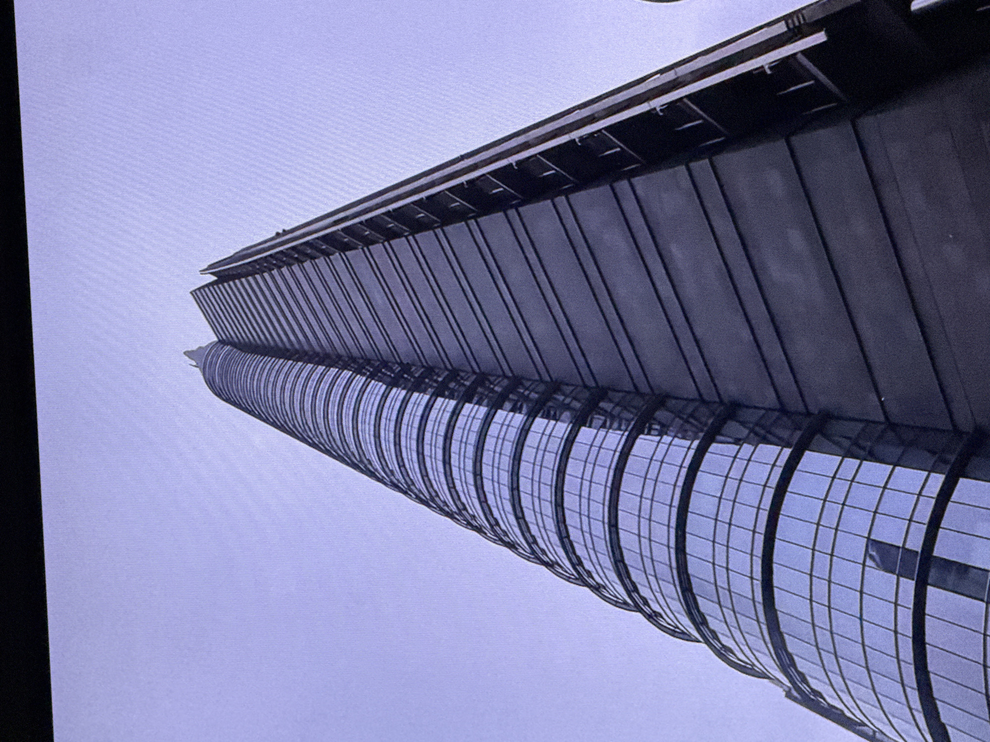 The media file shows a low-angle, tilted shot of a modern skyscraper, likely located in Dubai, United Arab Emirates. The building features a distinctive curved glass facade with a grid pattern, reflecting the overcast sky. Above the glass, the upper section of the skyscraper appears to be clad in dark, vertical panels, creating a contrasting texture. The overall scene suggests an urban environment characterized by contemporary architecture. The lighting and lack of harsh shadows indicate it is likely daytime, but the overcast conditions do not allow for a precise determination of the time of day. There are no people visible in the frame. The image focuses solely on the architectural elements of the building.