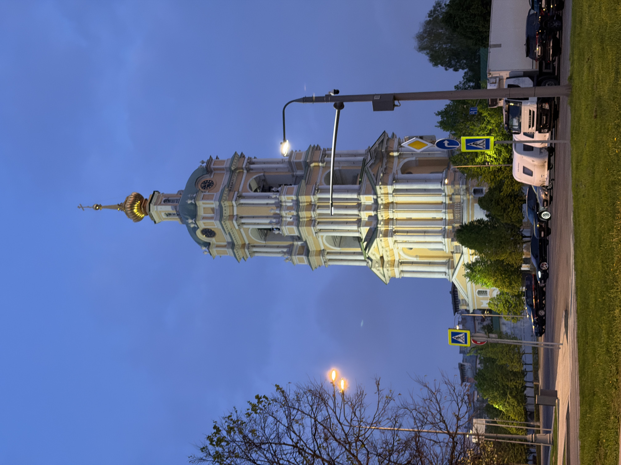 The image depicts the Church of the Intercession at Fili, located in Moscow, Russia. The baroque-style architecture of the church is prominent, illuminated against the backdrop of a darkening blue sky. Street lights provide additional illumination. Below the church, bare trees line the sidewalk. A row of vehicles is parked along the road. A pedestrian crosswalk with signage is visible.