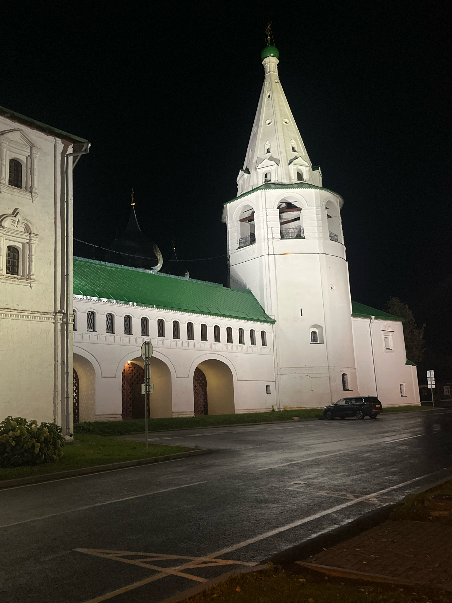 A night-time exterior shot features a white bell tower with a green spire, characteristic of Russian Orthodox architecture, which is illuminated against a dark sky. Adjacent to the bell tower is a long, white building with a green roof, featuring a series of arched entrances and arched windows. A dark-colored SUV is parked on the wet asphalt road in front of the building. The road shows reflections of streetlights, indicating it may have recently rained or is damp. The scene appears to be set in Suzdal, Russia, a city known for its historical churches and monasteries. The overall mood is serene and somewhat imposing, highlighted by the dramatic lighting and the historical architecture.