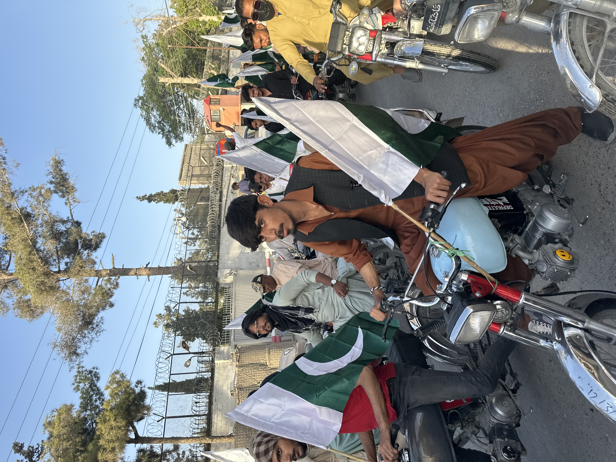 A street in Quetta City Tehsil, Pakistan, shows a gathering of people on motorcycles holding white and green Pakistani flags. Several people are riding motorcycles along the street. The scene appears to be a celebration or demonstration, based on the flags. The weather appears to be sunny and clear. A barbed-wire fence is visible.