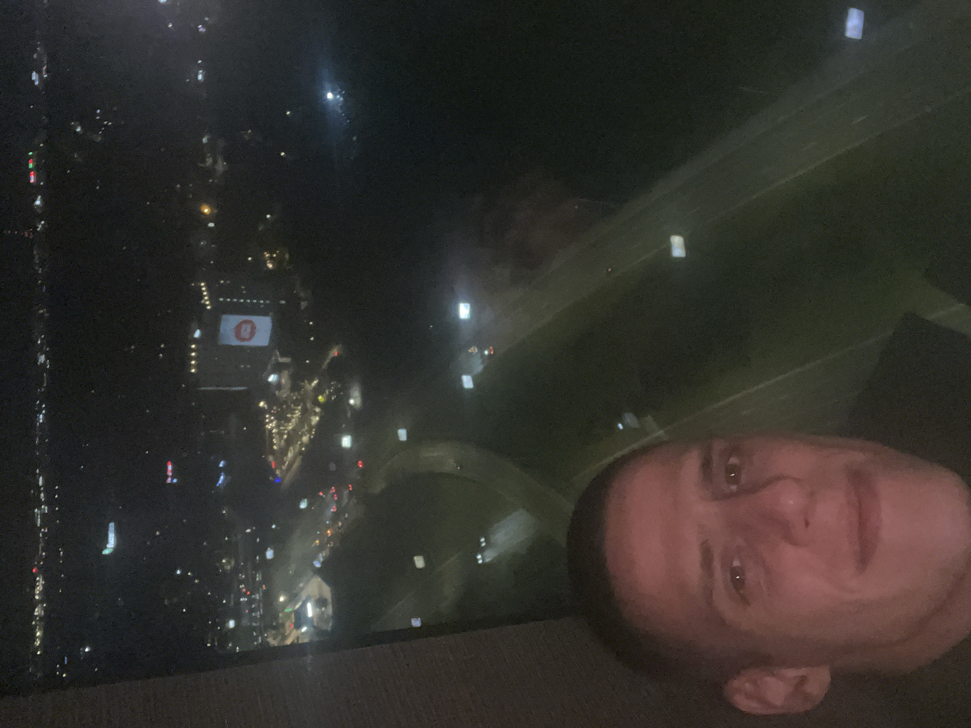 The image displays a night-time scene, oriented sideways, featuring a man's face in the foreground on the right, and a sprawling city view in the background on the left. The man's head is positioned such that he is looking forward and slightly upwards relative to his head's orientation, suggesting a selfie taken with the camera rotated. His features are visible, illuminated by an unseen light source that casts a warm glow on his skin, contrasting with the dark exterior.
The background reveals a panoramic view of an urban landscape at night, identified by the provided context as Sofia, Bulgaria. The dark sky is dotted with numerous artificial lights from buildings, streets, and vehicles, creating a vibrant, glowing mosaic across the left side of the frame. A prominent detail in the lower-middle portion of the city view is a brightly lit, rectangular sign on a building, displaying a distinctive red circular logo with a white internal symbol. Further into the distance, a wide, dark expanse traversed by what appears to be a multi-lane highway or major road is visible, marked by a series of square-shaped lights. The overall scene captures an elevated perspective of a bustling city under the cloak of night. There is no other discernible text beyond the logo.