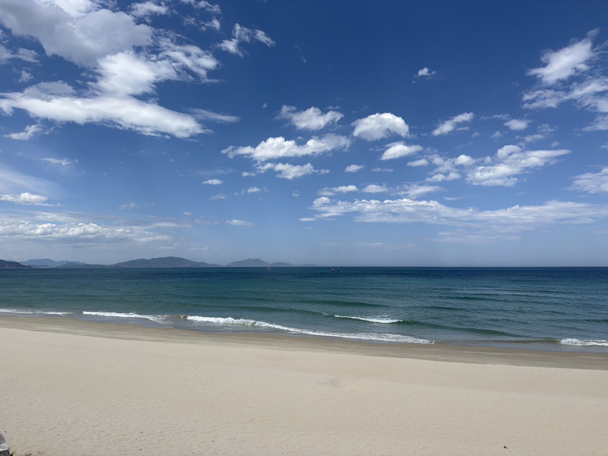 This image depicts a serene beach scene in Cam Duc, Vietnam. The foreground is dominated by a vast expanse of light-colored sand, gently sloping towards the ocean. The sand appears undisturbed, suggesting a quiet and tranquil atmosphere.
The middle ground showcases the turquoise-blue waters of the sea, with gentle waves rolling towards the shore. The water's surface is marked by subtle ripples, indicating a light breeze. In the distance, a thin line of green hills, characteristic of the Cam Duc landscape, stretches across the horizon. A few small boats are visible on the water, their presence subtle and not detracting from the overall peacefulness of the scene.
The sky above is a vibrant blue, dotted with scattered white cumulus clouds. The clouds are varied in shape and size, some appearing fluffy and others more wispy, adding depth and texture to the sky. The lighting suggests it is daytime, likely late morning or early afternoon, with the sun casting a bright, clear light on the entire scene. There are no people visible in the image, further emphasizing the sense of solitude and natural beauty. The overall impression is one of a calm, idyllic coastal environment.
