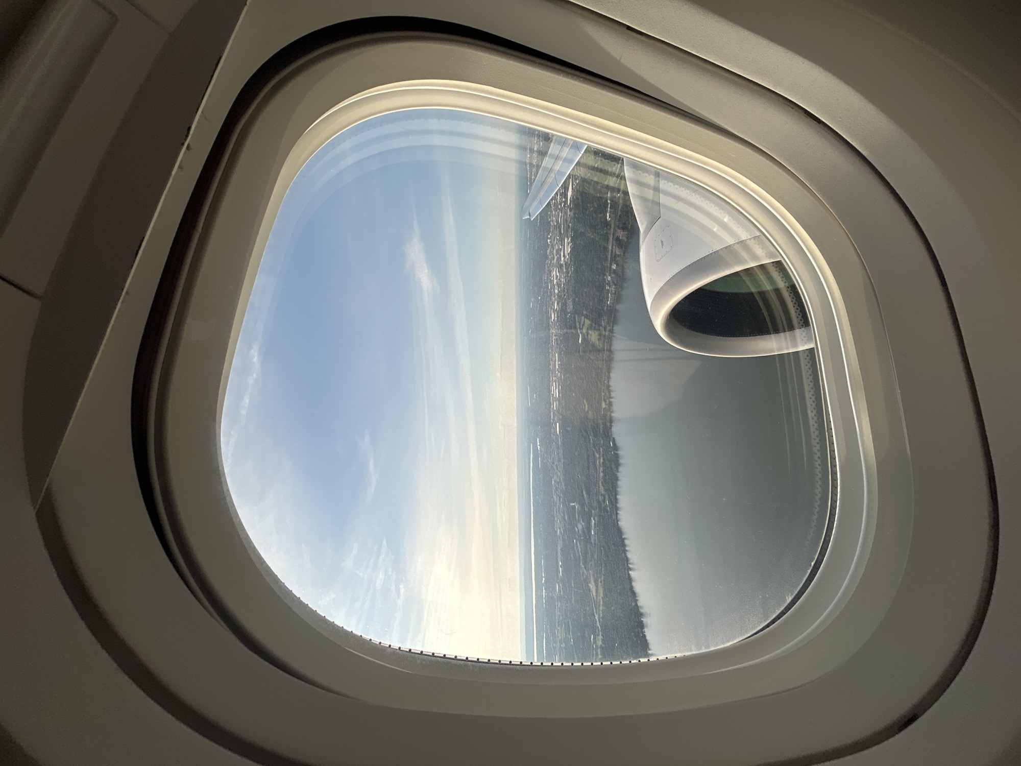 This image is taken from an airplane window, looking out at a landscape. The view through the oval window includes a large body of water in the foreground, with a treed shoreline. Beyond the water, a city or town stretches out, with buildings and infrastructure visible. An airplane engine nacelle is partially visible in the lower right corner of the window. The sky is blue with some wispy clouds. The interior of the airplane cabin is visible framing the window. The sunlight suggests it is daytime. There is no indication of the specific location within Pointe-Claire, Canada, or any specific details about people or actions.