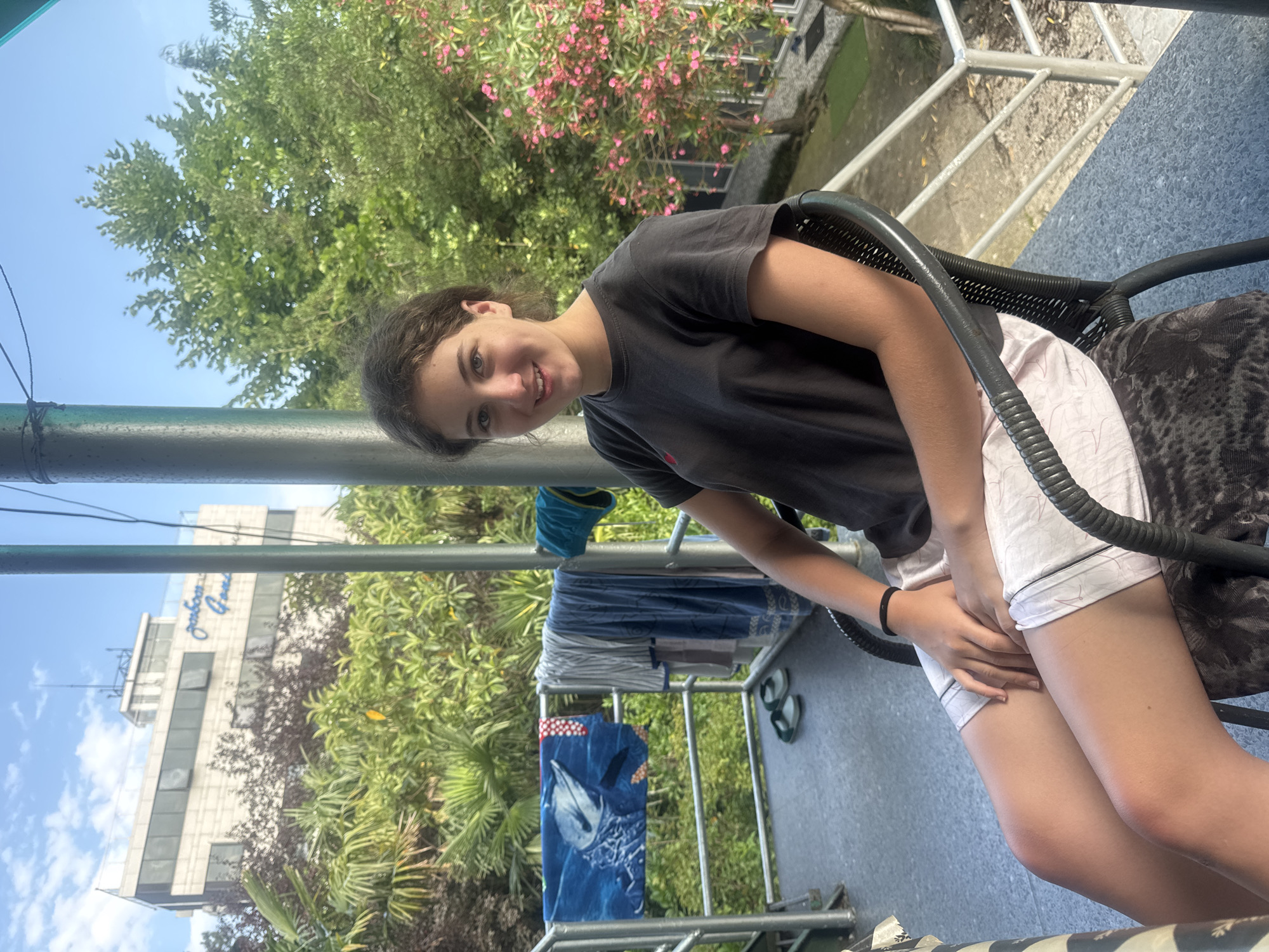 A young woman with curly brown hair and a light complexion is seated in a black wicker chair on an outdoor balcony. She is wearing a dark gray t-shirt with a small red logo and light-colored shorts. Her legs are crossed, and her hands are resting on her lap. She is smiling at the camera.
The balcony has a gray, speckled floor and a metal railing. On the railing, several towels are hanging to dry, including one with a dolphin print. A pair of green sandals is on the floor near the railing.
In the background, lush green foliage and trees are visible. Behind the trees, a white multi-story building with dark windows can be seen. The building has text on it in blue cursive script that reads "Gorbats Gorn".
The scene is lit by natural daylight, suggesting it is daytime. The overall impression is one of a relaxed outdoor setting.
Based on the building's signage "Gorbats Gorn", the location is identified as the Gorbats Gorn hotel in Batumi, Georgia.