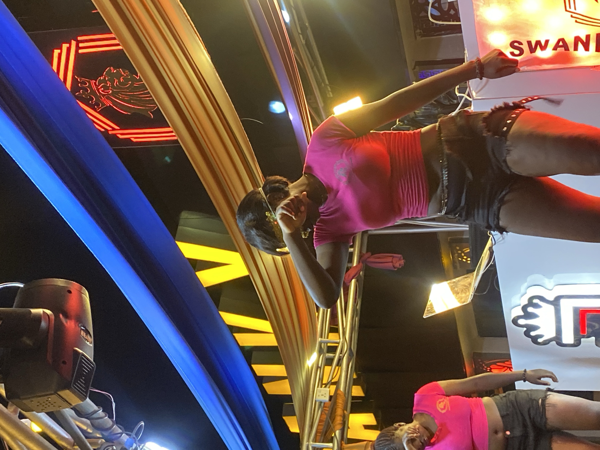 The image captures a nighttime scene at a venue in Bar Jirgi Summa, Nigeria, potentially a club or entertainment space. The primary subject is a young Black woman in the foreground, wearing a bright pink crop top and black distressed denim shorts. She is partially obscured by her own arm, which is raised to her face, suggesting she may be wiping her eyes or covering them. She also has a gold-colored headband on her forehead. To her left, another young Black woman, also in a pink crop top and black shorts, is partially visible. In the background, a DJ booth setup is present with a laptop. Above the performers, decorative elements include blue and gold fabric draped across the space, illuminated by yellow lights. A neon sign depicting a crowned lion's head is visible in the upper right portion of the image, along with red and yellow lighting. A professional-looking lighting rig is suspended above the area. The overall atmosphere appears to be festive and energetic.
