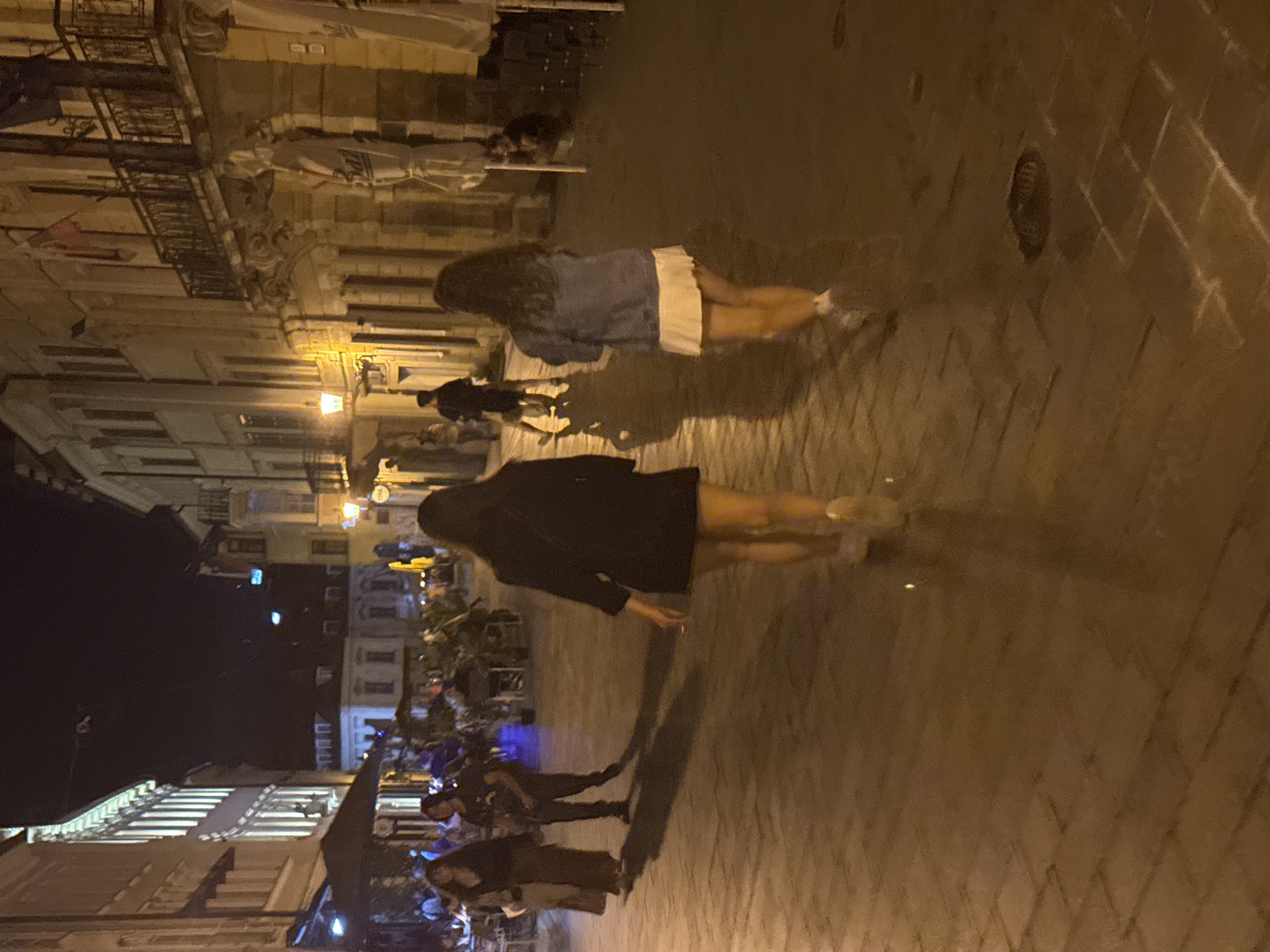 The image shows a night scene on a cobblestone street in Bratislava, Slovakia. Two women are walking away from the camera on the right side of the street. The woman in the foreground is wearing a dark jacket and a short skirt, and the woman behind her is wearing a denim jacket and a white skirt. Further down the street, two other individuals are walking in the same direction. On the left side of the street, several people are seated at tables outside a building, suggesting a restaurant or cafe. Buildings with illuminated windows line both sides of the street, casting a warm glow on the scene. The overall atmosphere appears lively and typical of a European city at night.