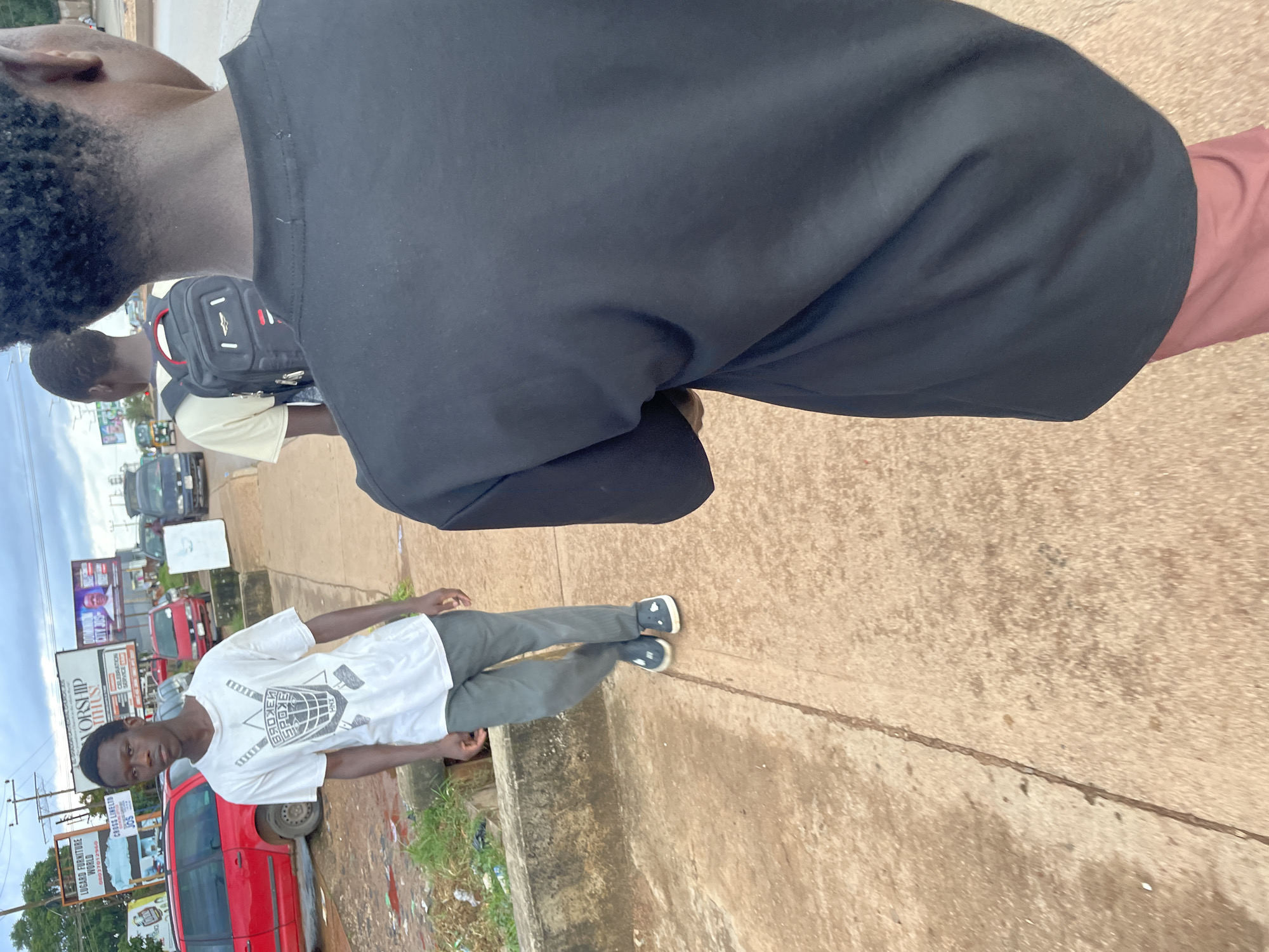 Two young Black males walk along a cracked concrete sidewalk next to a road in Jos, Nigeria. The male on the left, facing the camera, wears a white t-shirt with a shield-like graphic and gray pants. He has dark hair and a serious expression. The male on the right is seen from behind, wearing a black t-shirt and pink pants. He has a black backpack with red accents. The background shows a red vehicle, billboards, and other cars on the road. The sky is overcast. The scene appears to be a typical urban street in Jos.