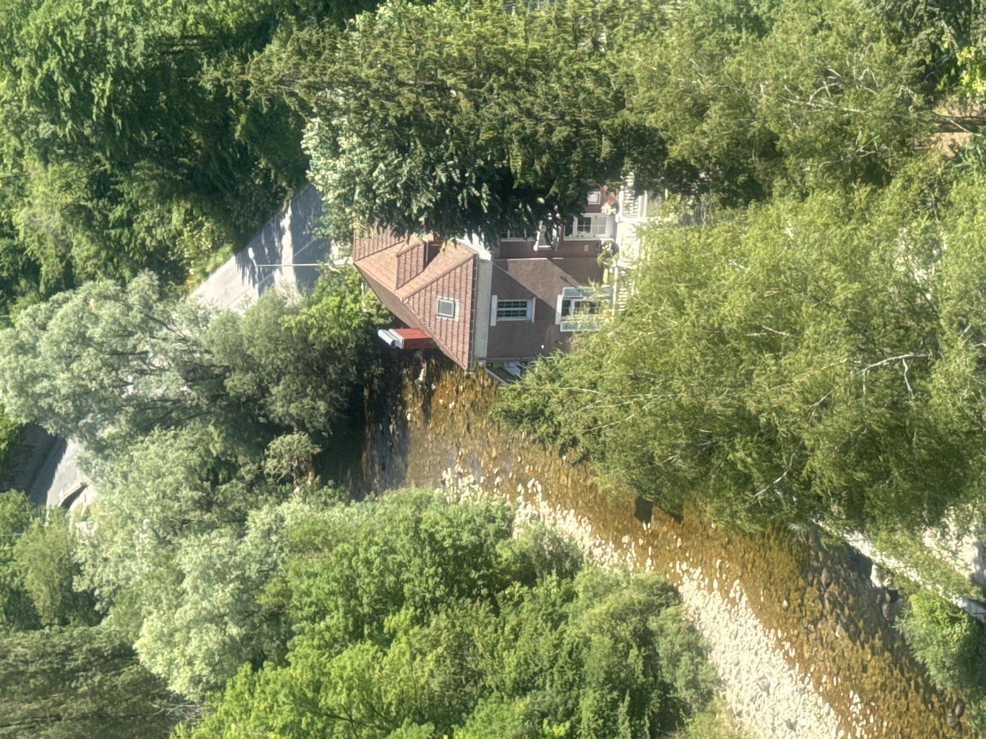 The image provides an aerial view of a winding road, a clear river with a stony bed, and a residential building nestled among dense green foliage in the city of Saint-Claude, France. The road, partially visible at the top of the frame, appears to be paved and slopes downwards. A lone streetlamp stands alongside the road. The river flows through a rocky channel, its water reflecting the surrounding greenery. On the riverbank, a multi-story house with a red tiled roof and visible windows is situated. The building features a chimney, a dormer window on the roof, and a porch area with white railings. Lush trees and vegetation dominate the landscape, creating a sense of seclusion and natural beauty around the property.