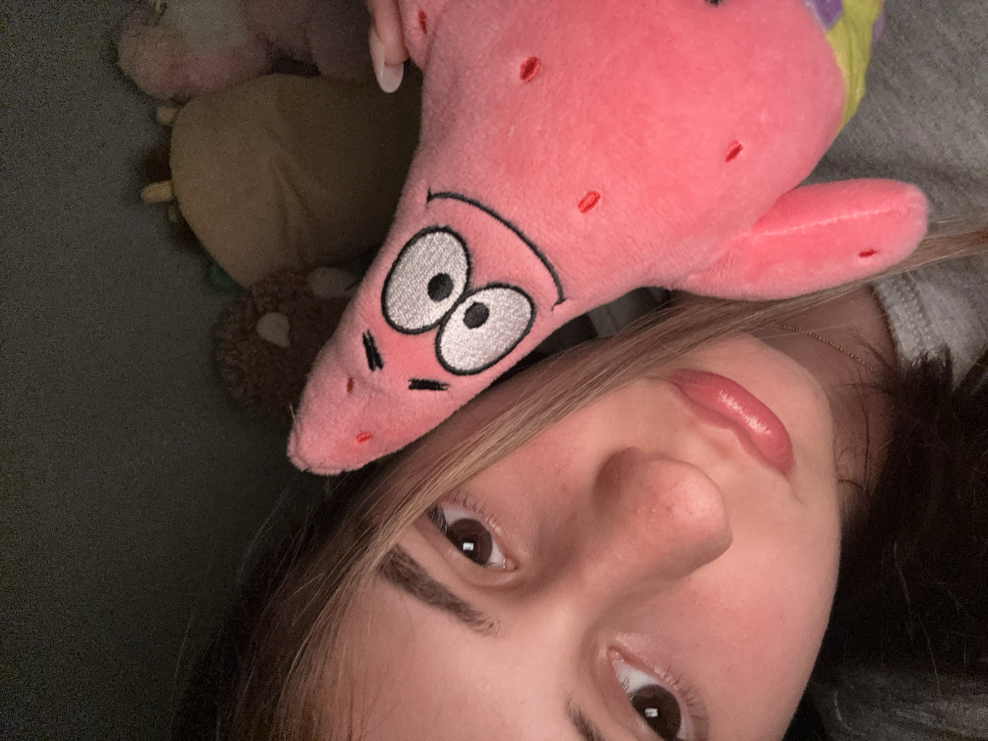 This close-up portrait shows a young person, likely female, lying down, with a pink plush toy of Patrick Star from SpongeBob SquarePants positioned on her head. The person has medium-toned skin, dark brown eyes that look directly at the viewer, and light brown hair with blonde highlights framing her face. Her lips are highlighted with glossy pink lipstick, and she has neatly defined eyebrows. Her expression is calm and neutral.
The Patrick Star plush is prominently featured, displaying its characteristic pink color with embroidered large white and black eyes, a curved black smiling mouth, and small red spots. A hint of yellow and purple fabric, likely part of Patrick's shorts, is visible near the top of the plush.
The background is dark and softly blurred, revealing other plush toys, including a brown one and a lighter, possibly tan, one, suggesting an indoor setting such as a bed or sofa. The soft, ambient lighting indicates an indoor environment, but no specific time of day is discernible. The overall mood of the image appears relaxed and playful. No visible text is present in the image. The location context for this media file is Panagyurishte, Bulgaria.