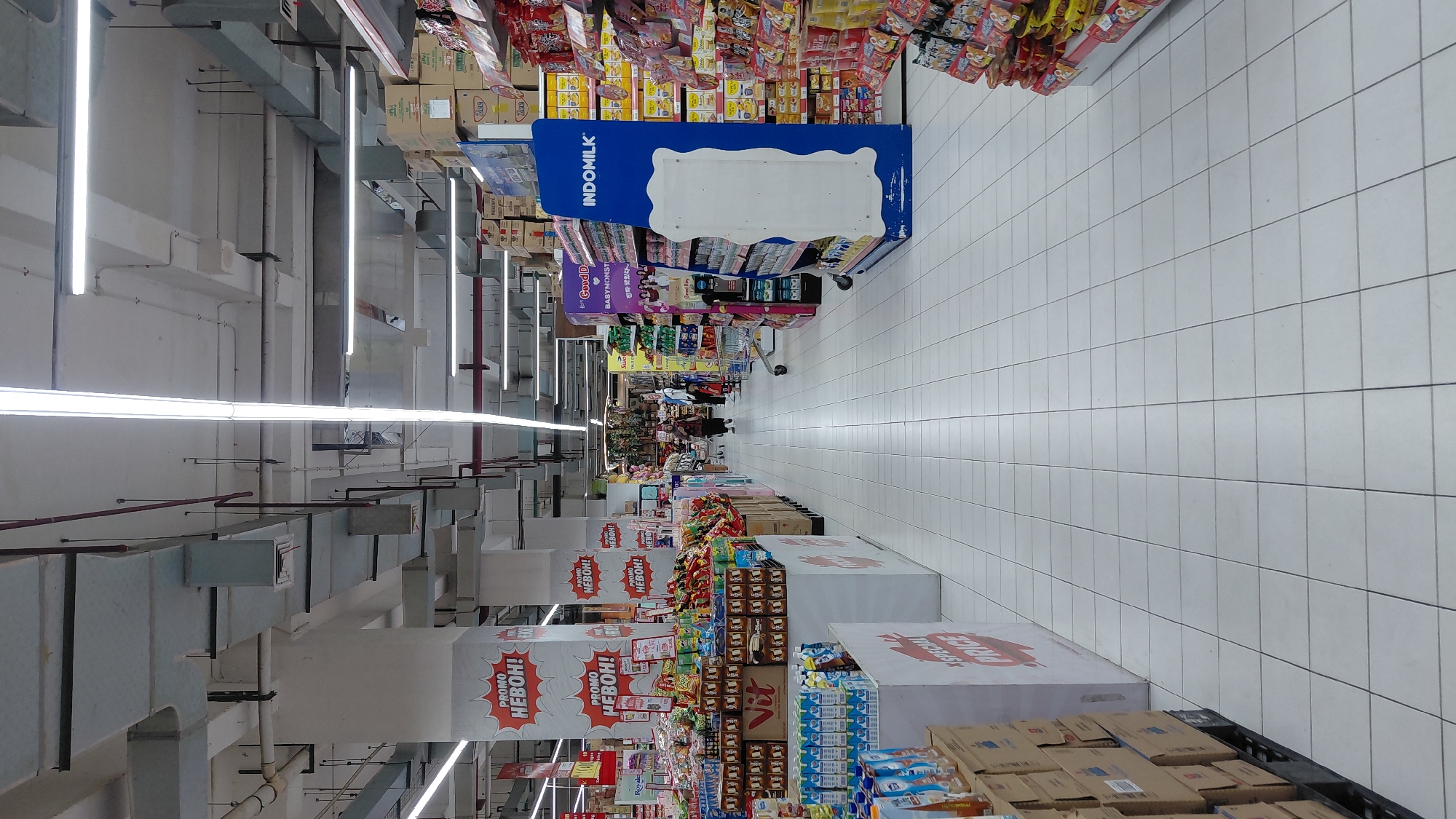 The image shows the interior of a well-lit supermarket aisle in Purwokerto, Indonesia. The floor is tiled in white squares, and fluorescent lights run along the ceiling. On the left side of the aisle, there are shelves stocked with beverages, boxed goods, and promotional displays with text in Indonesian, including "PROMO HEBOH!". On the right side, there are more shelves displaying packaged snacks and food items, with a prominent blue display stand labeled "INDOMILK". Two individuals, one adult and one child, are visible walking down the aisle in the distance. The overall environment suggests a typical retail setting.