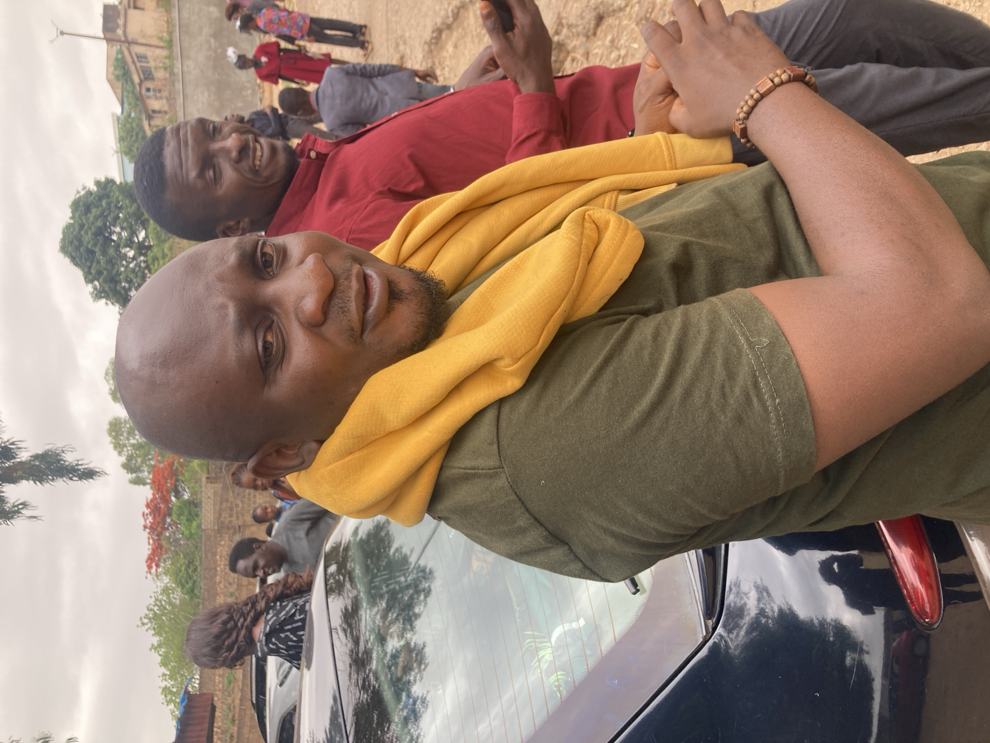 The image captures two men in an outdoor setting. One man, closer to the camera, is wearing an olive-green t-shirt with a yellow hoodie draped around his neck. He appears to be reclining. The second man, slightly behind and to the side, is wearing a red button-down shirt and grey pants. He is looking towards the camera and smiling. In the background, there are several other individuals, along with trees and buildings. The rear windshield of a dark-colored car is visible in the foreground. The presence of a specific building structure matches the architectural style found in the city of Jos, Nigeria, and given the landscape, it is likely that the photo was taken in the city of Jos, Nigeria.