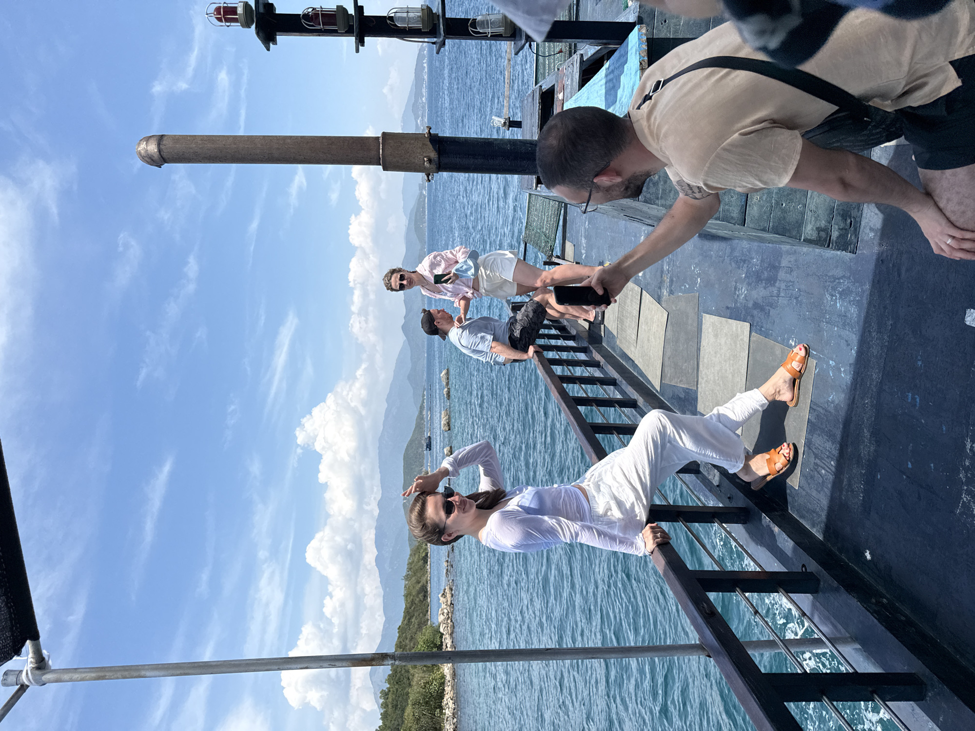 Captured in Nha Trang Ward, Vietnam, the scene depicts a group of four individuals aboard a vessel sailing on the sea. A woman in white clothing and brown sandals is lounging against the railing, her left arm shielding her eyes from the sun. Behind her, two men and a woman stand closer to the mast. One man is crouched down, extending his hand towards the railing. The environment consists of the sea and the land, with cloud-filled sky.