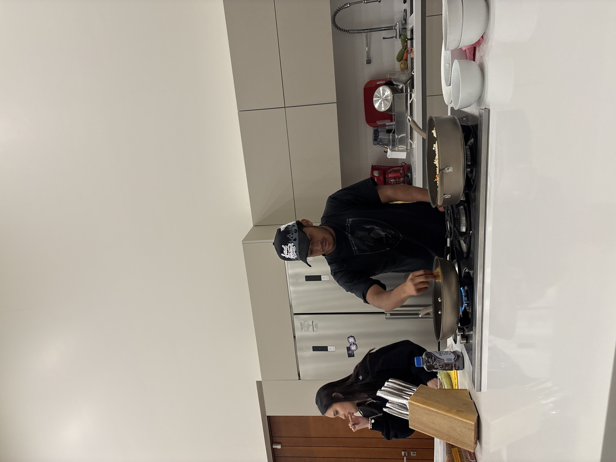 A man wearing a black t-shirt and a black cap is cooking in a modern kitchen. He is holding a large pan over a lit gas stovetop. Next to him, a woman in black is standing at the counter. A knife block and a water bottle sit on the counter nearby. Behind them is a refrigerator. The location is Santiago Etla, Mexico.