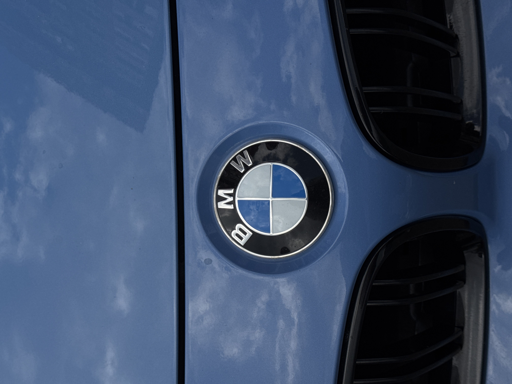 A close-up, eye-level shot captures the front emblem of a blue BMW car. The emblem is circular, with a silver outer ring and a black inner ring. Inside, it features four quadrants: two are light blue and two are grey. The letters "B", "M", and "W" are visible in silver against the black background. The car's blue paint reflects the cloudy sky. Below the emblem, a portion of the car's black grille is visible. The image is a static shot, with no people or actions depicted, and no specific location in Krasnodar, Russia can be identified from the visual information provided.