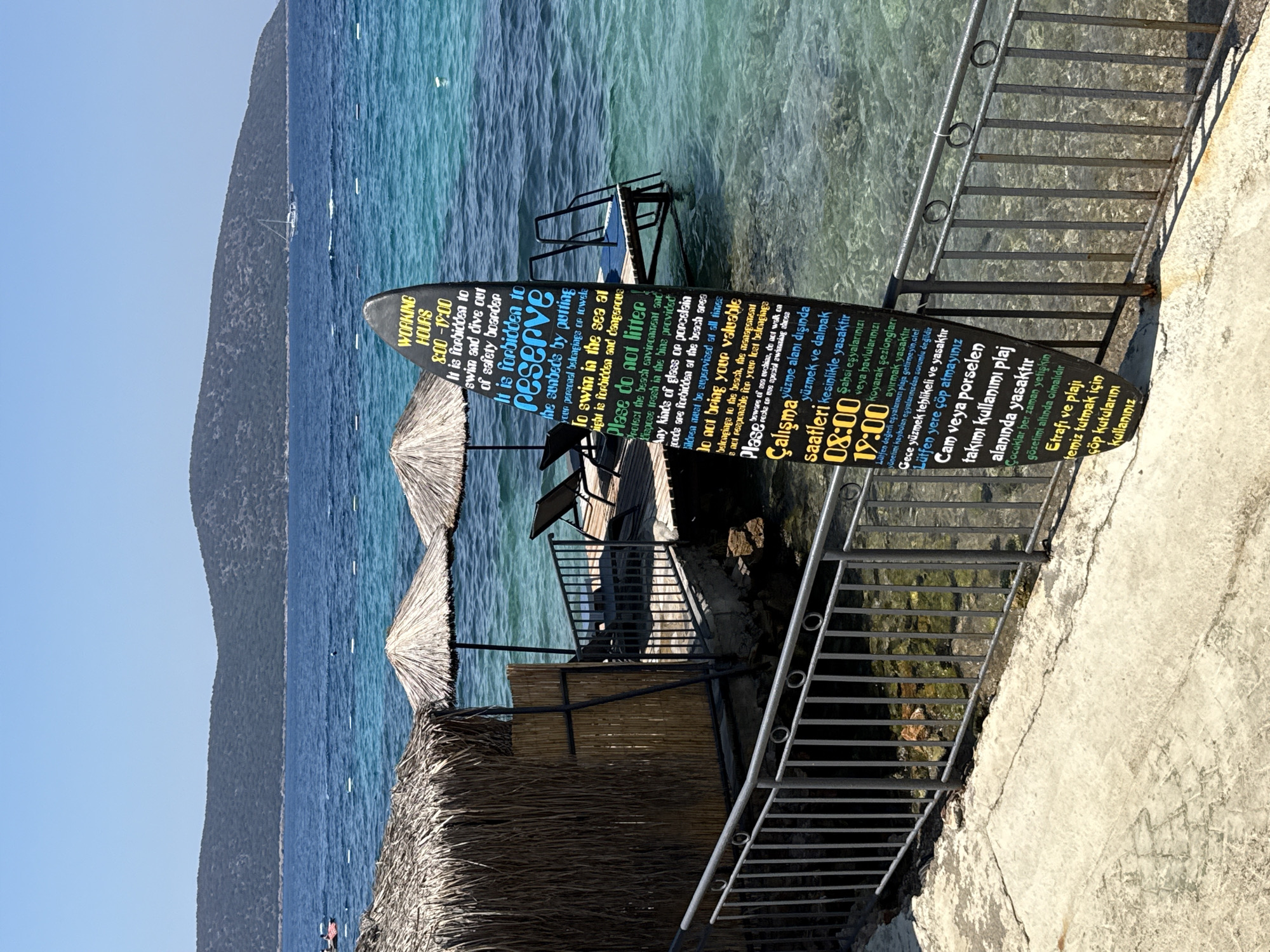 The image depicts a coastal scene in Bodrum, Turkey, characterized by clear turquoise water and a mountainous backdrop. In the foreground, a concrete walkway runs alongside a metal railing, overlooking the sea. To the left, a rustic structure with a thatched roof and woven walls provides shade. Beyond this, a wooden jetty extends into the water, equipped with loungers and a ladder for accessing the sea.
The central element of the scene is a surfboard-shaped sign standing upright on the beach. The sign displays rules and information in both English and Turkish, indicating operating hours from 8:00 to 19:00. It prohibits swimming and diving outside of safety boundaries and discourages reserving sunbeds with personal belongings. The sign also advises against littering, warns about the dangers of swimming in the sea, and states that the management is not responsible for lost valuables. Additionally, it prohibits glass or porcelain items, advises against bringing valuable items to the beach, and notes that children must be supervised. The Turkish portion of the sign reiterates these rules, including prohibitions on swimming outside designated areas, keeping personal items, and the operating hours.
The sea is calm with visible buoys marking an area. In the distance, a sailboat is visible on the horizon. The overall impression is one of a well-maintained beach area offering recreational activities and clear guidelines for visitors.