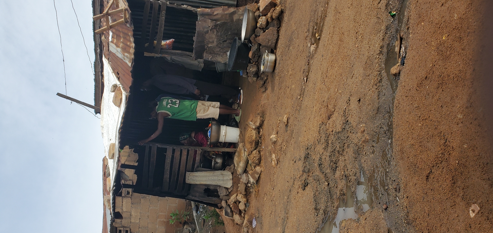 In an outdoor scene of a dilapidated structure, two men and a woman are present. The structure is made of corrugated metal and wood, with a makeshift roof of white tarp and tin. In front of the structure, the ground is dirt and gravel, with puddles of water in uneven terrain. The man on the left, wearing a green basketball jersey with "JAMES 23" on the back, stands near the entrance of the structure, leaning with his left arm on the wooden frame. The woman, seated inside the structure, is wearing a reddish-pink garment and a headscarf. To the right of the man in the jersey, another man in a dark shirt and dark pants stands with his back to the camera. Several objects are scattered around the entrance, including large metal bowls, buckets, and rocks. A white plastic bucket is positioned near the woman, and a large black basin is visible to the right, with another metal basin on top of rocks. The background shows a brick wall and more of the corrugated metal structure. The sky is overcast.