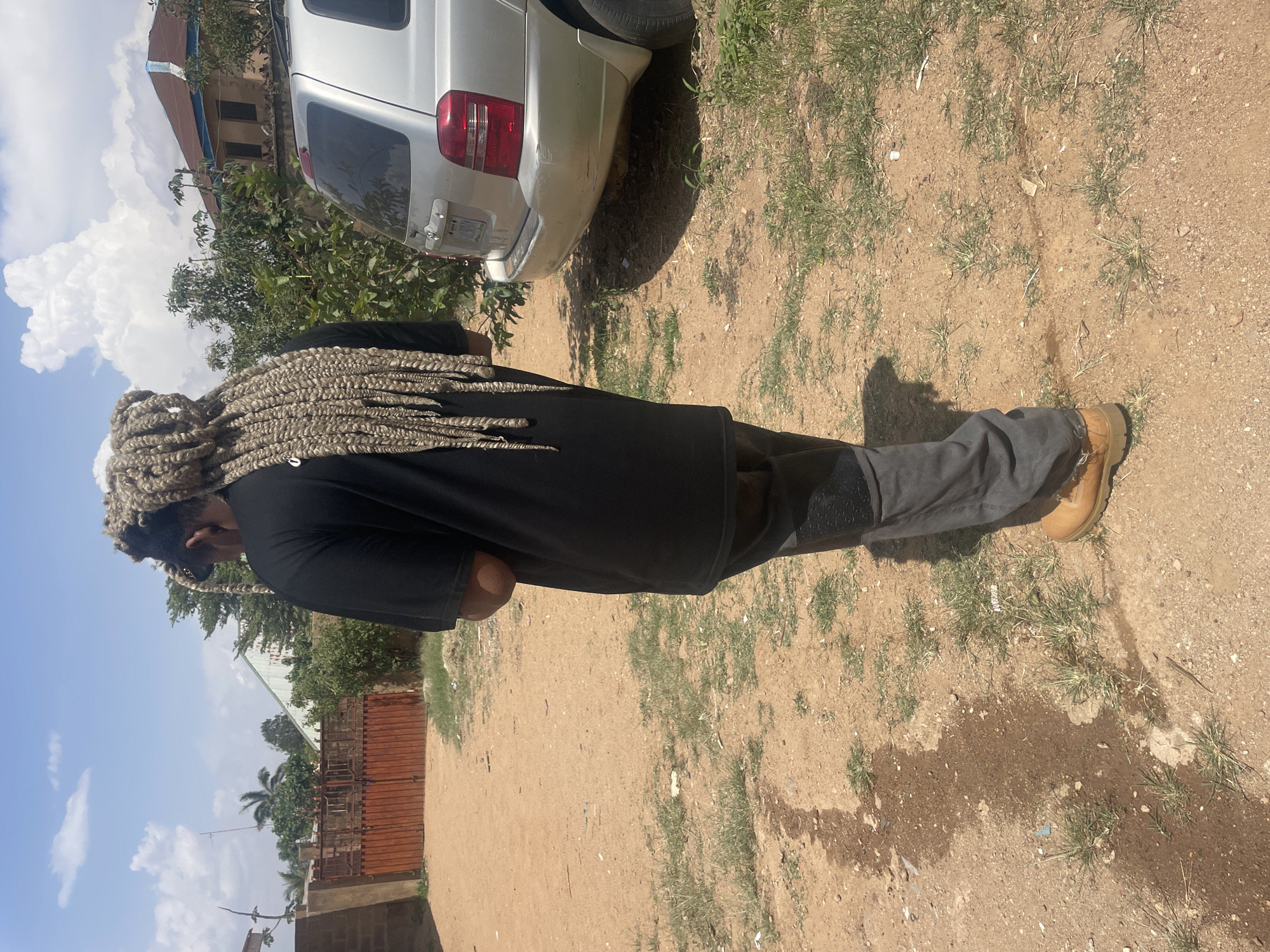 The photo shows a person standing in a dirt lot beside a silver SUV. The individual is wearing a black shirt, grey pants, and tan boots. They have long, light-colored braids. The ground is dirt with patches of grass. In the background, there is a fence, trees, buildings, and a blue sky with clouds. Considering the features, the precise location within Jos, Nigeria, is not discernable from the image alone.