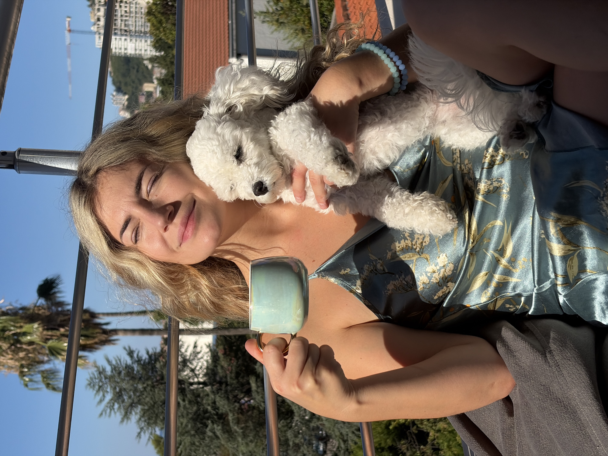 A young woman with blonde, wavy hair is holding a small white poodle while sitting on a balcony overlooking Budva, Montenegro. She is wearing a teal silk slip dress with a floral pattern and has a light blue mug with a gold handle in her left hand. The dog is nestled in her arms, and she has her eyes closed, appearing relaxed. In the background, palm trees, a dense green forest, and buildings can be seen, with construction cranes visible in the distance, indicating a city setting. The sky is clear and blue, and the scene is bathed in bright sunlight. A grey cushion is visible to her left, and she is wearing two light blue beaded bracelets on her right wrist.