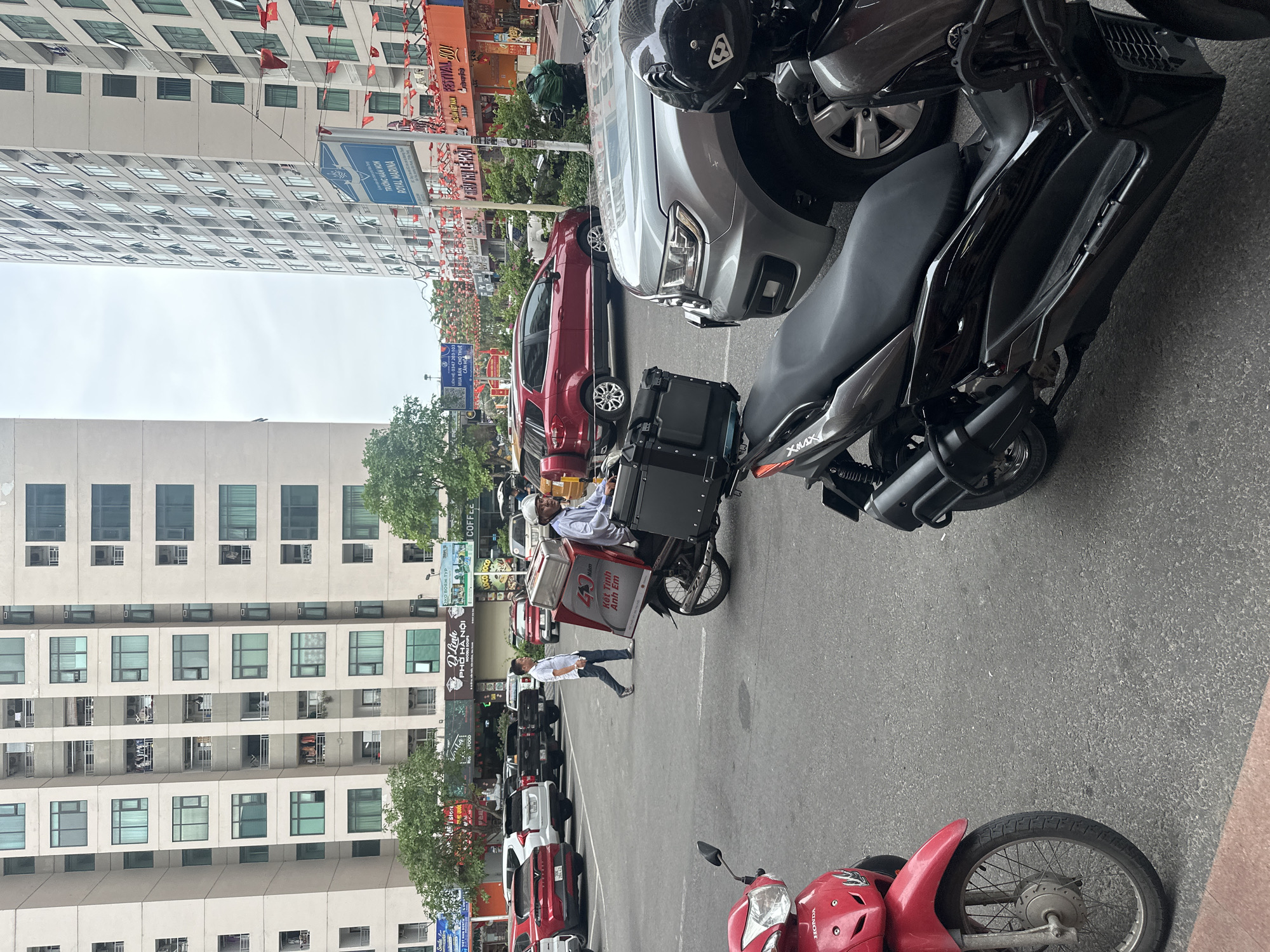 The photograph captures a street scene in Bac Nha Trang Ward, Vietnam, showing a parking area along a street lined with buildings. Parked cars and motorbikes fill the scene. A man, possibly a delivery driver, stands beside a parked motorcycle fitted with a delivery box. The delivery box is red and white, and appears to have the words "Ket Tinh Anh Em" on it. Further back, a man in a white shirt is seen walking on the street. Buildings flank the street, including a tall apartment building on the left. Street signs and banners adorn the street, including ads for "Royal Marina" and "Festival 100!" A sign for "D'Linh Pho Ha Noi" can also be seen. Vietnamese flags are strung across the street. The parking area is gray, suggesting a paved surface. The sky overhead is overcast.