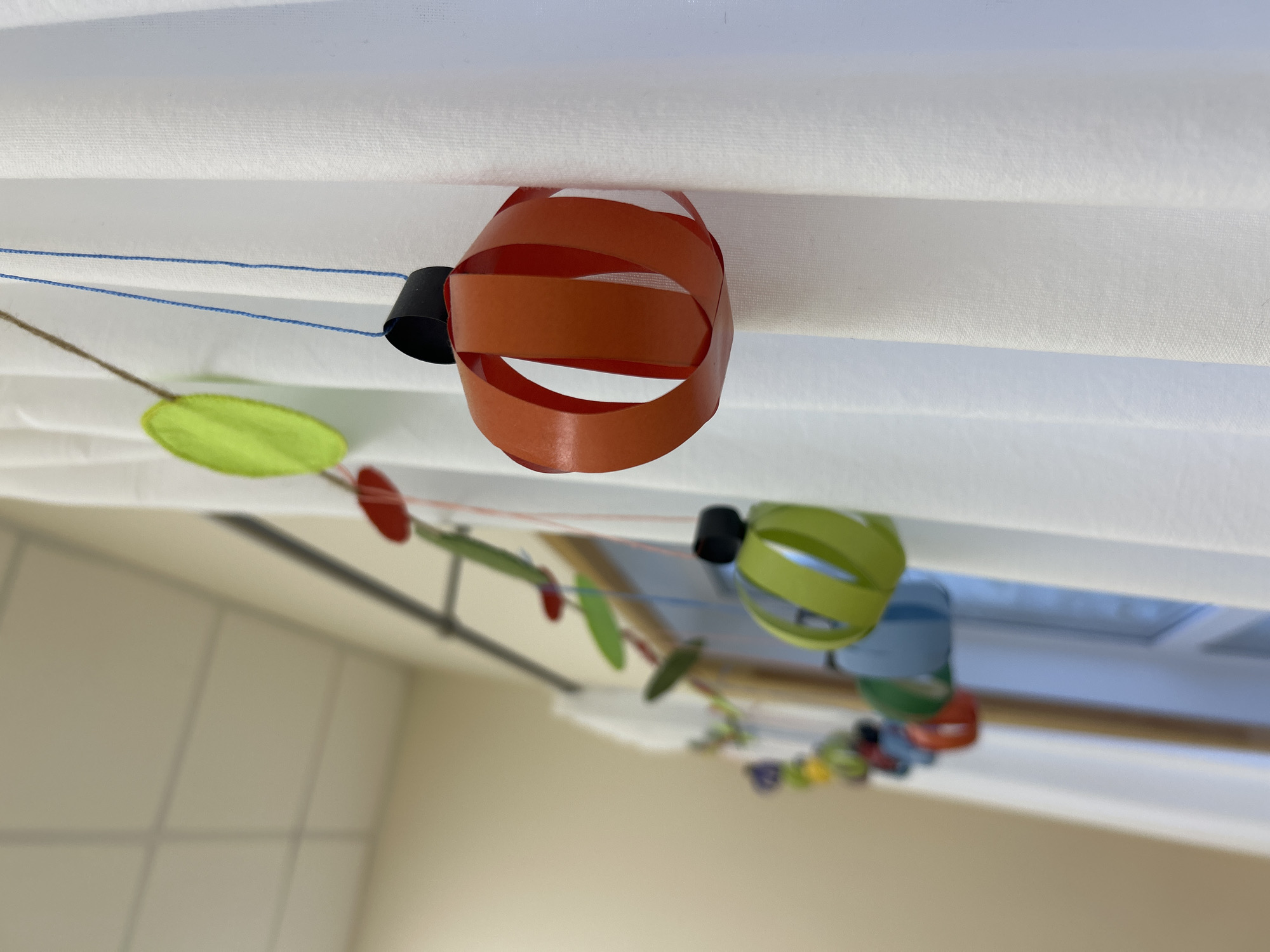 The image shows a string of paper decorations hanging indoors, likely from a ceiling. The decorations are circular, made from strips of colorful paper. The colors include orange, green, blue, red, yellow. The decorations are attached to a string, held in place by small black cylindrical pieces. The background includes a white surface (possibly a ceiling or blind) and a blurry view of a wall and window. There is no information to suggest the location is Izhevsk, Russia.