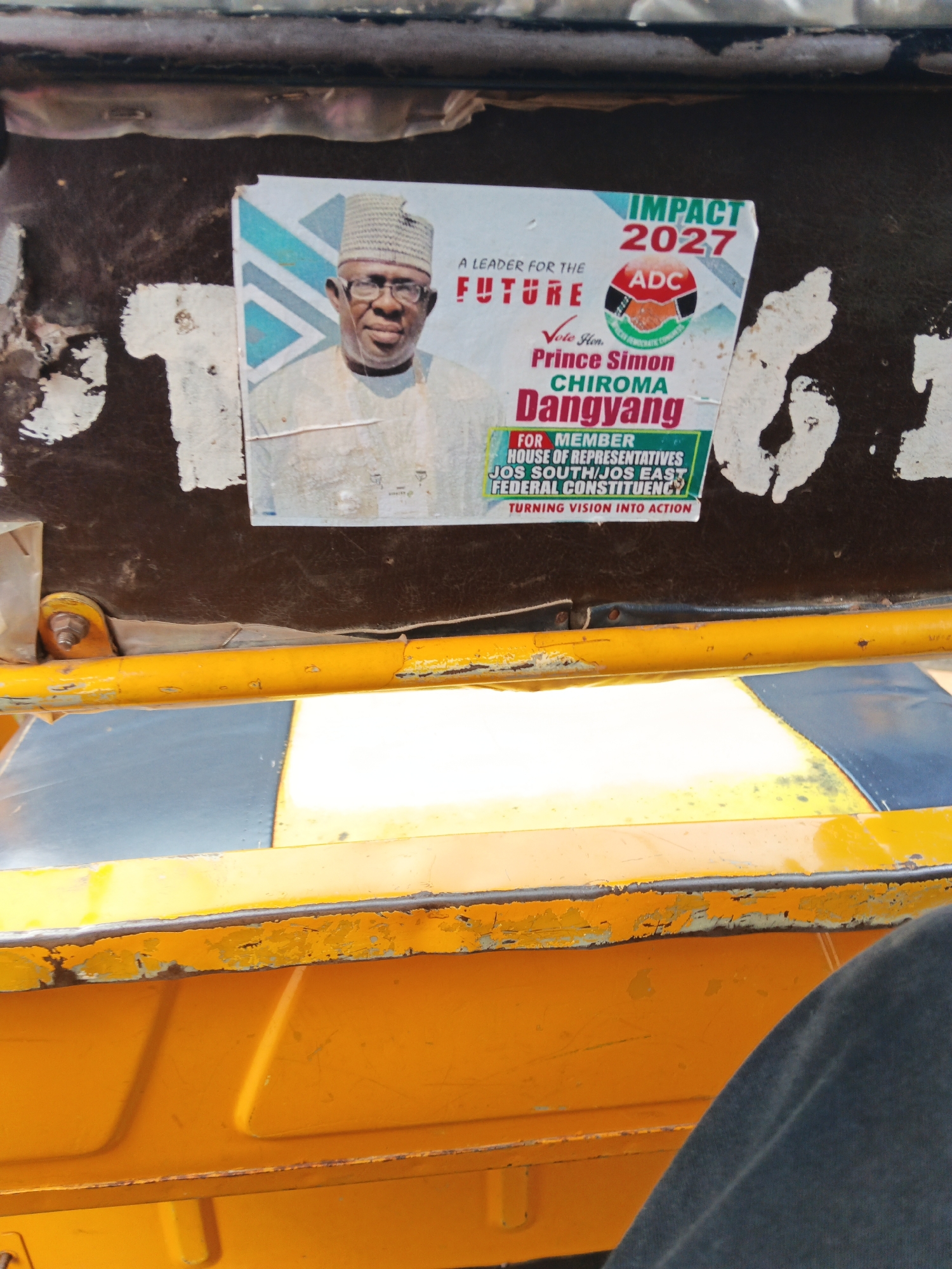 The image shows a close-up view of a poster stuck on the dark brown interior of a vehicle, likely a rickshaw or a similar public transport. The poster features a portrait of a man wearing glasses and a patterned cap. He is dressed in light-colored traditional attire. Text on the poster indicates it's a campaign poster for "Prince Simon Chiroma Dangyang" for the position of "Member House of Representatives Jos South/Jos East Federal Constituency." It also includes the slogan "IMPACT 2027" and the logo of a political party, "ADC." Below the portrait, there are phrases like "A LEADER FOR THE FUTURE" and "TURNING VISION INTO ACTION."
The poster itself is slightly worn and peeling at the edges, suggesting it has been exposed to the elements. The background of the poster has a geometric pattern in shades of blue and white. The vehicle's interior appears to be made of dark, textured material, with some scuff marks and remnants of old stickers visible. A bright yellow metal bar runs horizontally across the lower portion of the image, with black and grey striped panels underneath it, which might be part of the vehicle's body or seating. The lighting suggests it is daytime, with natural light illuminating the scene. The image is taken from a low angle, looking upwards towards the poster.