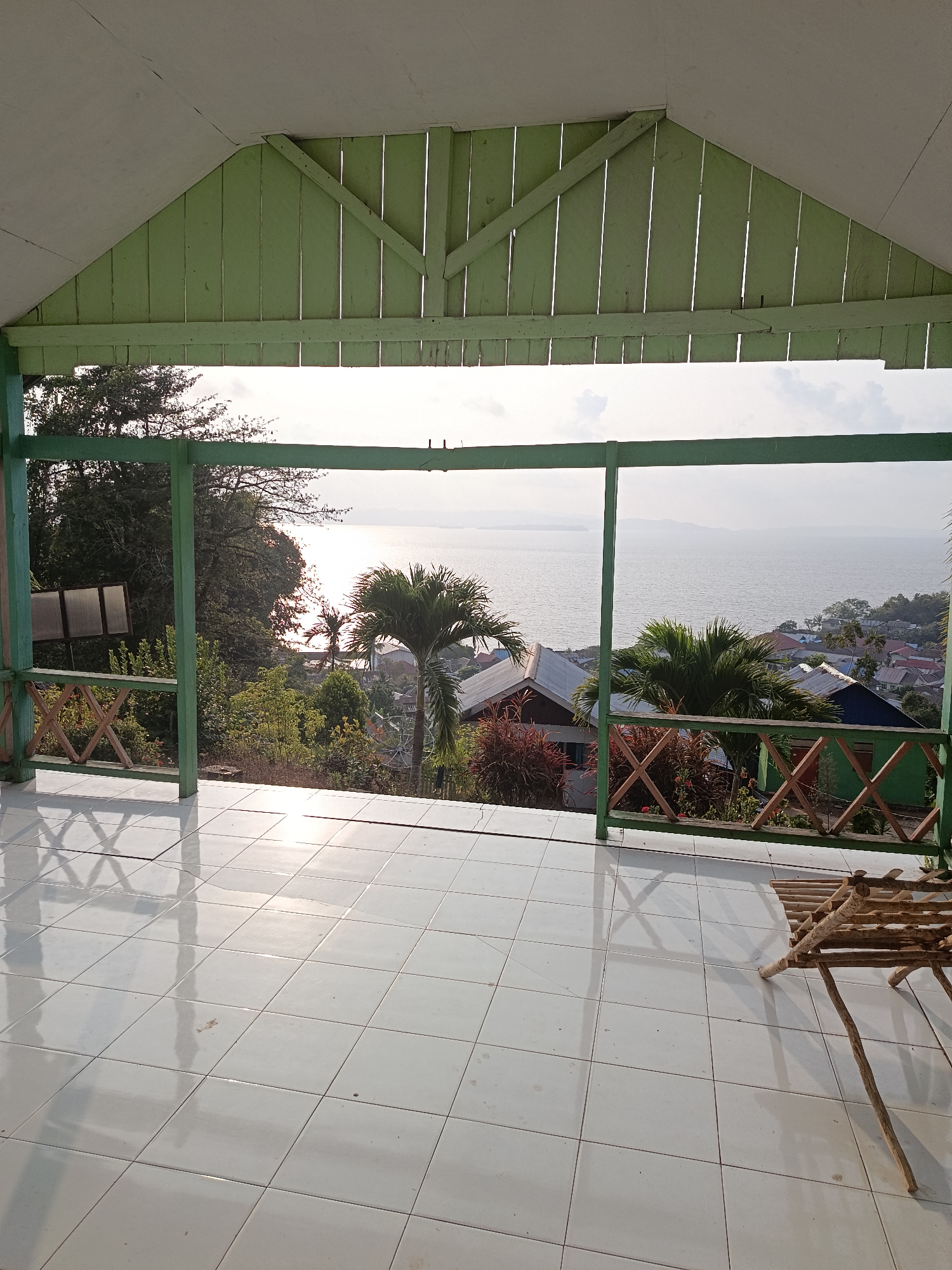 This is an eye-level, medium outdoor shot taken from a covered porch area overlooking a coastal town in Mansalean, Indonesia. The foreground features a white tiled floor with some shadows cast from the porch railings and a small, rustic wooden chair. The porch is framed by light green painted wooden pillars and railings with a lattice design. Beyond the porch, a lush green landscape slopes down towards a body of water. The water, likely a bay or ocean, reflects the bright, hazy sunlight, creating a shimmering effect. In the distance, a town with various houses, many with red or blue roofs, is visible nestled among the trees and along the coastline. Palm trees are scattered throughout the scene, including a prominent one in the mid-ground. The sky is overcast with some diffused sunlight. The overall impression is one of a serene, tropical view from a residential vantage point.