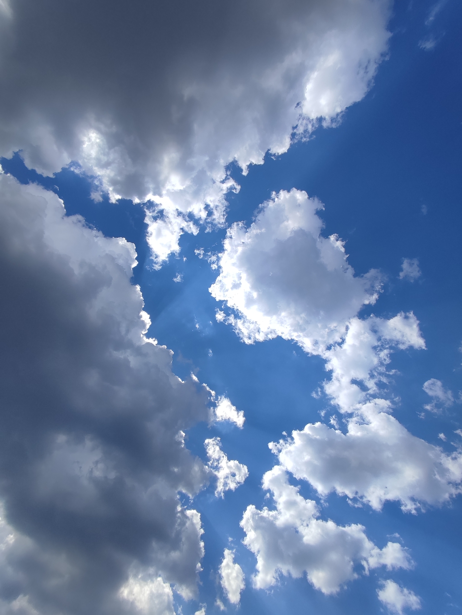 The image shows a sky with a mix of dark gray and white cumulus clouds, with patches of clear blue sky visible between them. Sunlight is filtering through the clouds in several places, creating rays of light. The location is not determinable from the visual information.