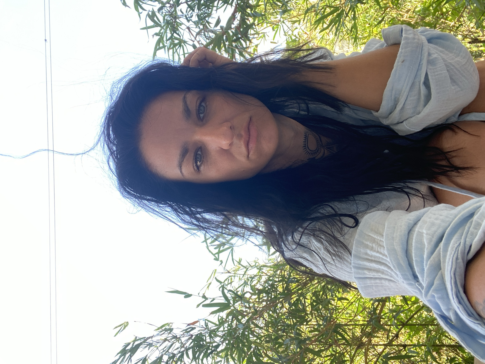 A medium shot depicts a woman with dark hair and green eyes, possibly of European descent, set against a background of lush green vegetation, likely bamboo. She is wearing a light blue button-down shirt. A visible tattoo adorns her neck. The location is likely in Mũi Né, Vietnam, based on the presence of bamboo, a common plant in that region. A single, thin wire or cable is visible extending from the upper left of the frame towards the top, likely a utility line.